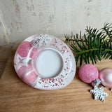Rosa Teelichthalter aus klarem Epoxidharz, ca. 8 × 6,5 cm, Höhe ca. 4 cm. Im Harz sind kleine rosa Weihnachtskugeln, ein Stern, eine Schneeflocke und rosa Granulat eingegossen. Ovaler Halter für ein Teelicht, Unterseite mit vier runden Korkpads als Möbelschoner. Fotografiert auf Holz mit Tannenzweig und rosa Kugeln als weihnachtlicher Dekohintergrund.