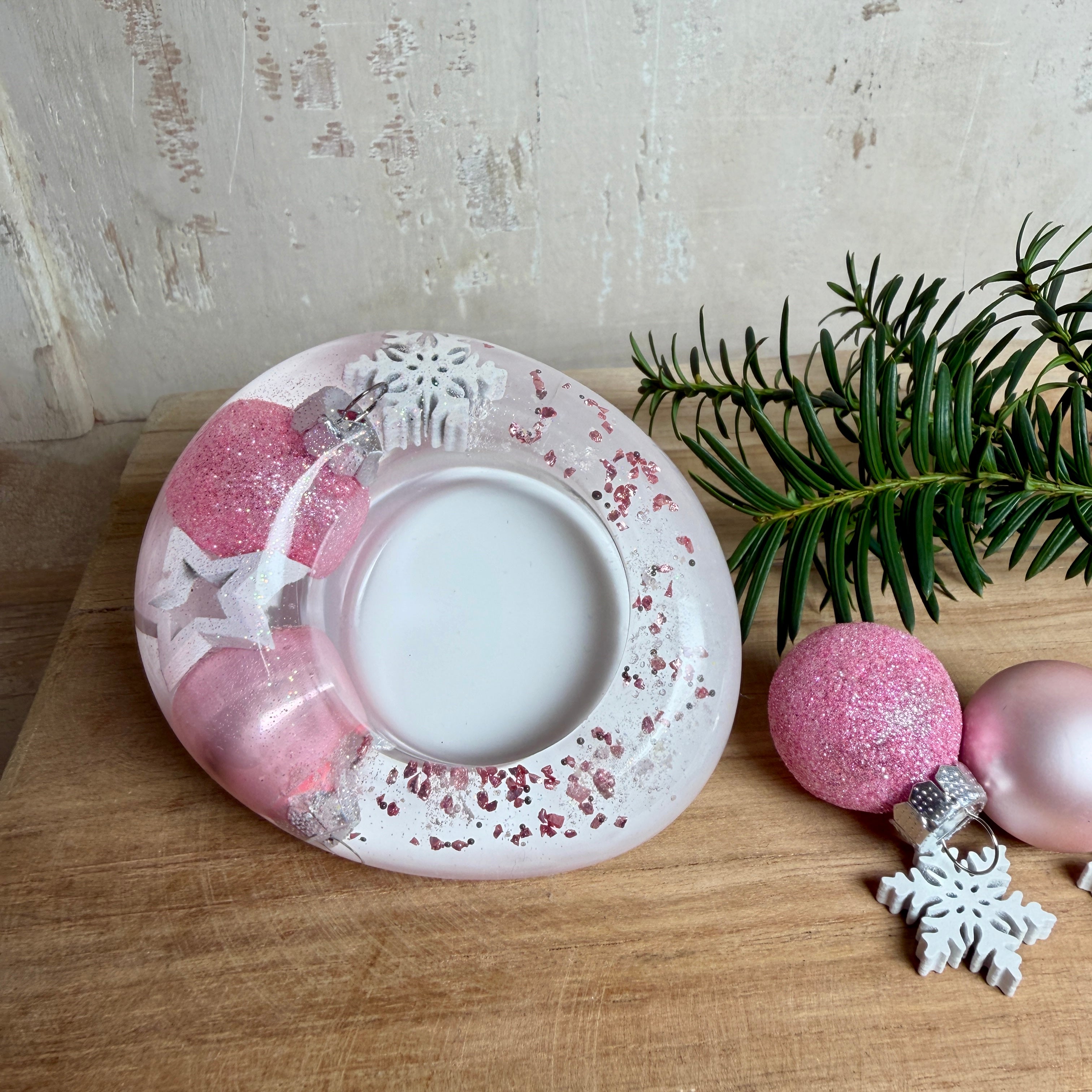 Rosa Teelichthalter aus klarem Epoxidharz, ca. 8 × 6,5 cm, Höhe ca. 4 cm. Im Harz sind kleine rosa Weihnachtskugeln, ein Stern, eine Schneeflocke und rosa Granulat eingegossen. Ovaler Halter für ein Teelicht, Unterseite mit vier runden Korkpads als Möbelschoner. Fotografiert auf Holz mit Tannenzweig und rosa Kugeln als weihnachtlicher Dekohintergrund.