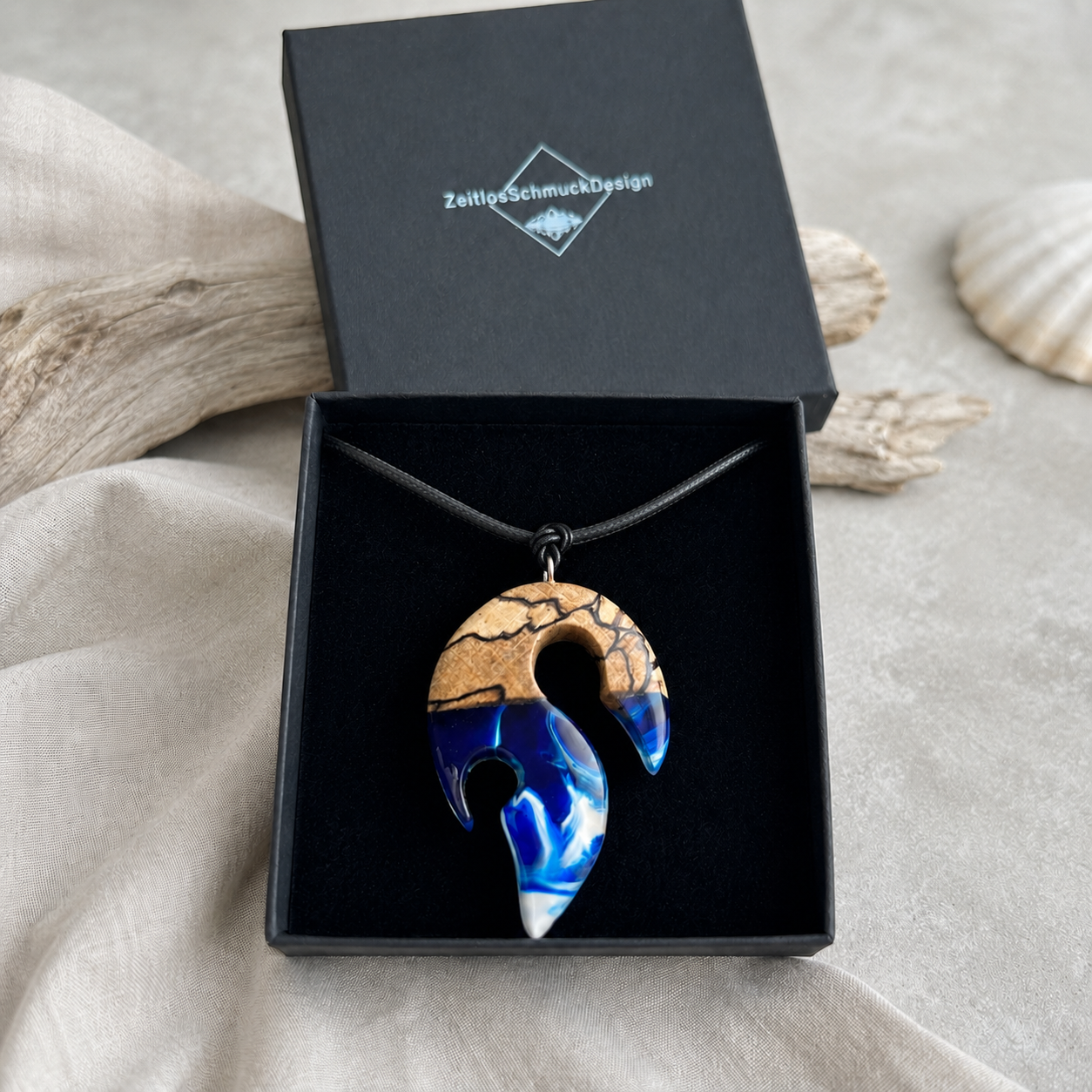 Schmuckbox-Bild TIDE – handgefertigter Holz-Harz-Anhänger aus Buchenholz und blau-weißem Epoxidharz in Maori-Angelhaken-Form, am schwarzen Lederband in schwarzer Schmuckbox von ZeitlosSchmuckDesign präsentiert.