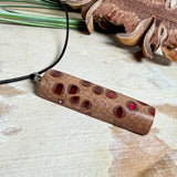 Handgefertigter Halsanhänger aus echtem Banksiazapfen, veredelt mit rotem Harz in den natürlichen Kammern; ca. 5,5 × 1,6 cm. Mit Anhängerschlaufe aus Edelstahl an schwarzer Lederkette (45 cm + 5 cm Verlängerung) mit Edelstahl-Karabiner. Detailaufnahme bzw. Tragebild vor neutralem, natürlichem Hintergrund.