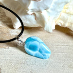 Larimar Freeform-Anhänger auf schwarzer Lederkette. Beidseitig tragbarer Trommelstein, ca. 2 x 1,8 cm groß, mit Silberöse. Der seltene, kostbare Larimar zeigt lebendige Blau- und Weißnuancen wie karibisches Lagunenwasser. Lederkette 45 cm mit Edelstahl-Karabiner und 5 cm Edelstahl-Verlängerung, fotografiert vor einer großen Muschel im maritimen Stil.