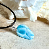 Larimar Freeform-Anhänger auf schwarzer Lederkette. Beidseitig tragbarer Trommelstein, ca. 2 x 1,8 cm groß, mit Silberöse. Der seltene, kostbare Larimar zeigt lebendige Blau- und Weißnuancen wie karibisches Lagunenwasser. Lederkette 45 cm mit Edelstahl-Karabiner und 5 cm Edelstahl-Verlängerung, fotografiert vor einer großen Muschel im maritimen Stil.