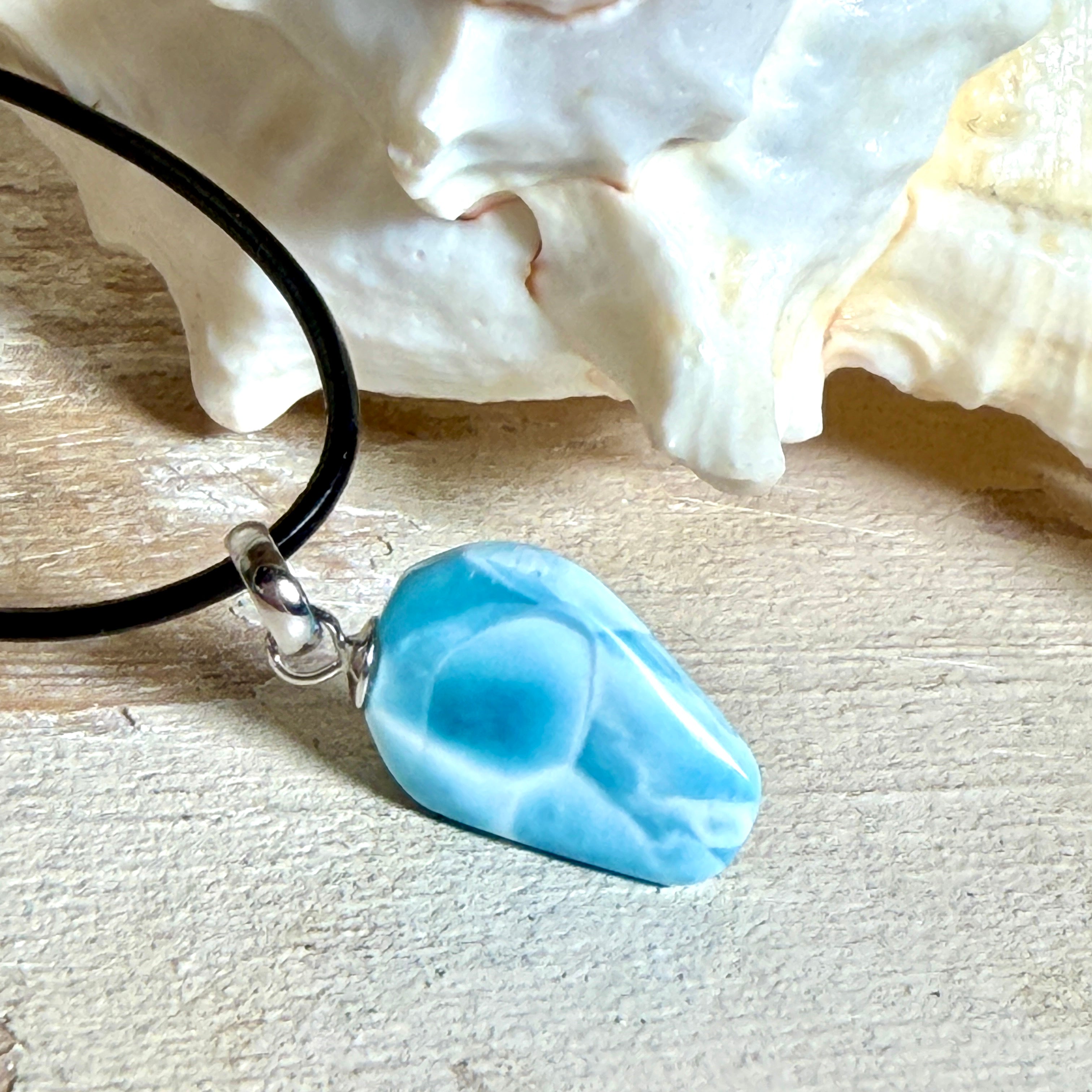 Larimar Freeform-Anhänger auf schwarzer Lederkette. Beidseitig tragbarer Trommelstein, ca. 2 x 1,8 cm groß, mit Silberöse. Der seltene, kostbare Larimar zeigt lebendige Blau- und Weißnuancen wie karibisches Lagunenwasser. Lederkette 45 cm mit Edelstahl-Karabiner und 5 cm Edelstahl-Verlängerung, fotografiert vor einer großen Muschel im maritimen Stil.