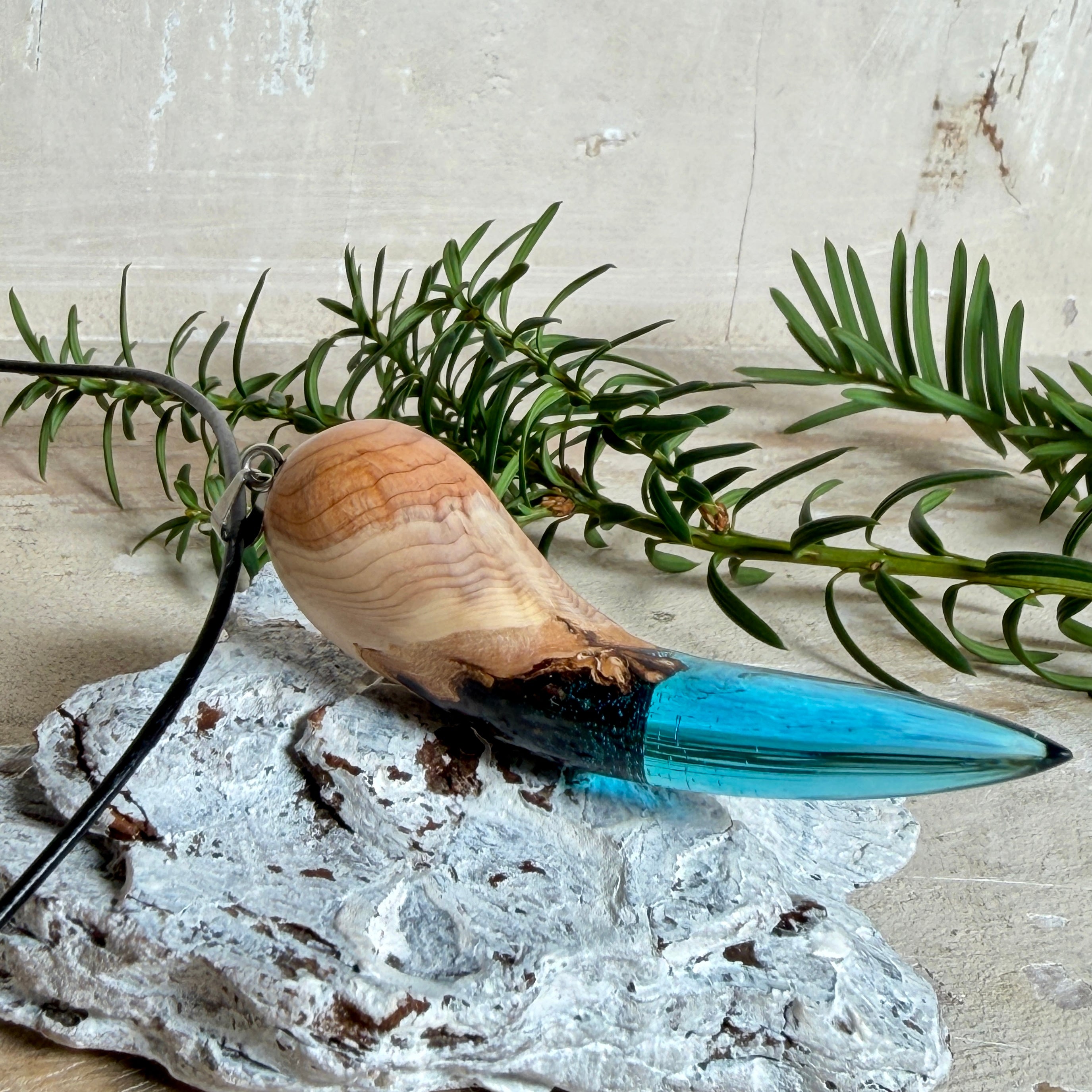 Handgefertigter Anhänger aus Edelholz und transparent-türkisblauem Epoxidharz, glatt poliert. Größe ca. 6,7 × 2,0 cm. Mit Edelstahlöse an schwarzer Lederkette (45 cm + 5 cm Edelstahl-Verlängerung, Karabiner). Maritimer, moderner Unisex-Schmuck mit natürlicher Holzmaserung; kleine Lufteinschlüsse sind charakteristisch.