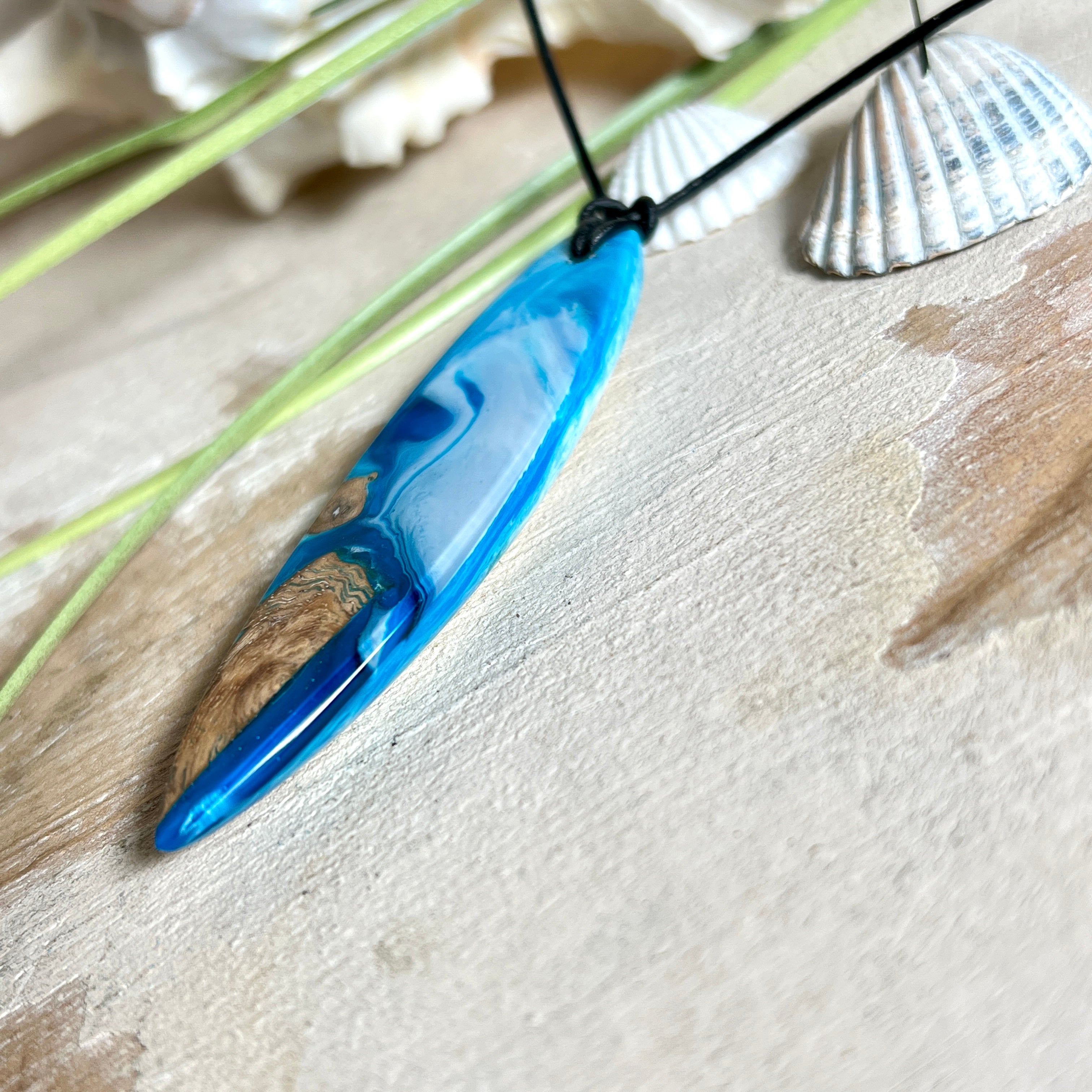 Handgefertigter Surfbrett-Anhänger „Ocean Glide“ aus Maserholz und Epoxidharz in verschiedenen Blau-, und Weißtönen. Der ca. 6,5 × 2 cm große Anhänger wird an einem verstellbaren Lederband (26–46 cm) mit Holzperlen geliefert. Ein maritimes Unikat mit natürlicher Holzmaserung, das die Schönheit des Meeres einfängt.