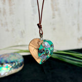 Herzförmiger Anhänger aus Olivenholz mit aquamarinfarbenem Harz und eingelegten Paua-Muschelstücken (Abalone), irisierender Blau-Grün-Schimmer. Der Anhänger hängt an einem braunen, verstellbaren Lederband mit Schiebeknoten; Aufhängung aus Edelstahl. Produktfoto vor maritimer Deko/Abalone-Schale.