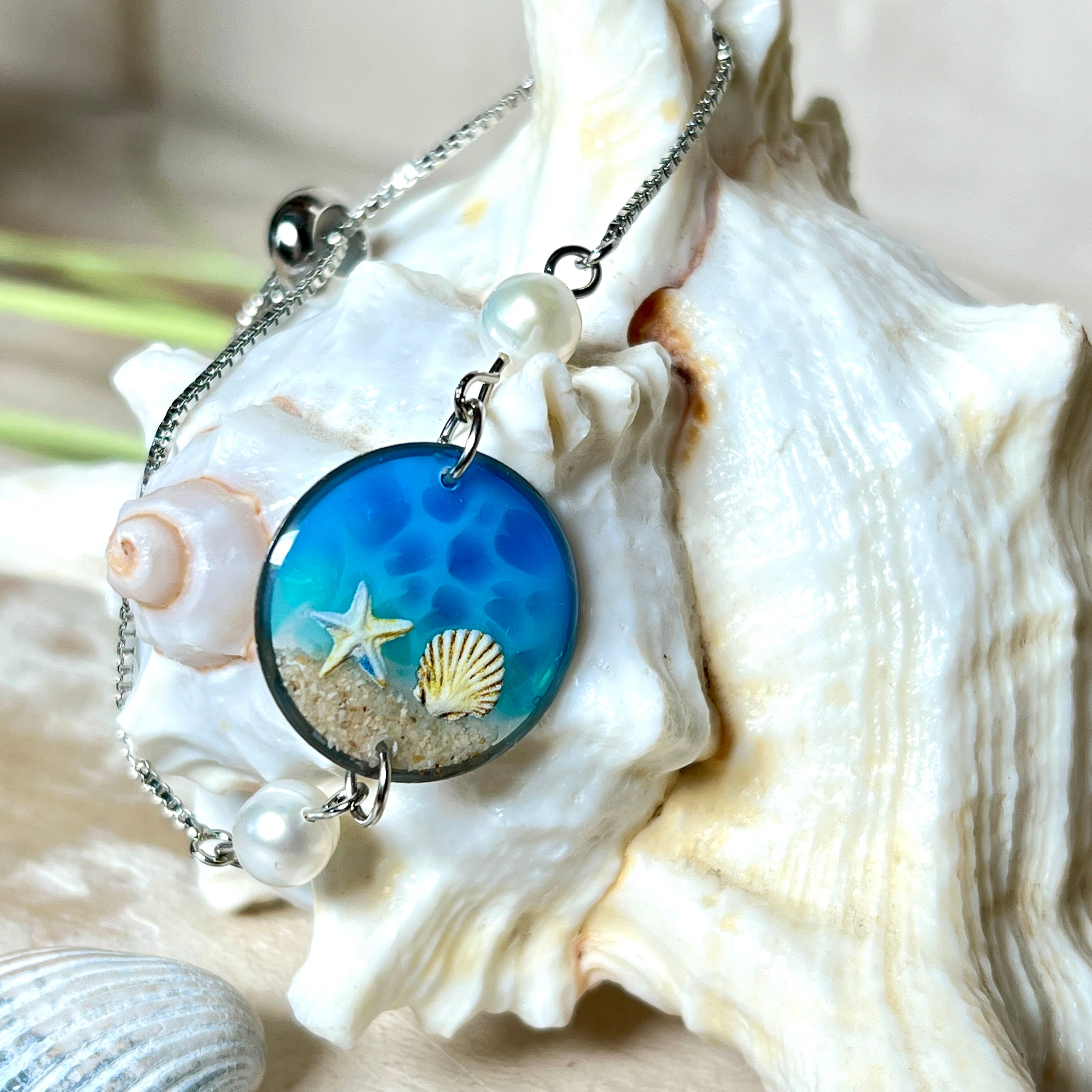 Elegantes Edelstahl-Armband mit rundem Resin-Anhänger in Meeresblau. Im Anhänger liegt echter Meersand als Ufersaum; darüber Sticker-Motive Seestern & Muschel. Zwei feine Süßwasserperlen verzieren das Armband. Verstellbarer Schiebeverschluss . Handgefertigtes Unikat – maritimer Schmuck, der an Wellenrauschen und Strandspaziergänge erinnert.