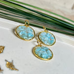 Runder Anhänger „Golden Atlantis Dream“ mit kleinen Larimar-Edelsteinchen in klarem Resin, gefasst in einer 24K vergoldeten Zinklegierung in Shiny Gold. Die Larimarsteine zeigen sanfte Blau- und Weißtöne wie karibische Wellen. Durchmesser ca. 22 mm, handgefertigtes Unikat, fotografiert mit Larimarsteinchen, Muscheln und einer 2-Euro-Münze zur Größenorientierung auf maritimem Untergrund.