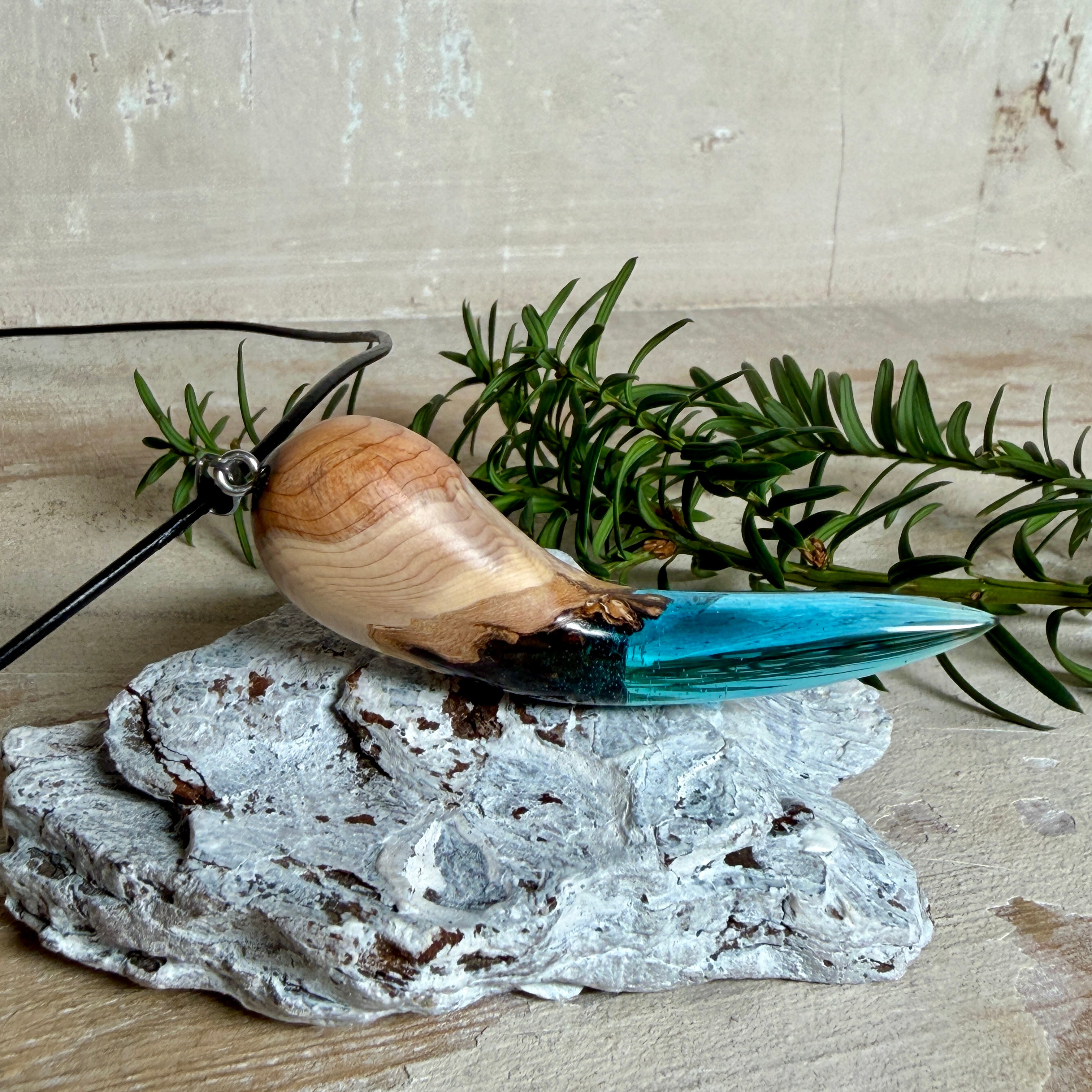 Handgefertigter Anhänger aus Edelholz und transparent-türkisblauem Epoxidharz, glatt poliert. Größe ca. 6,7 × 2,0 cm. Mit Edelstahlöse an schwarzer Lederkette (45 cm + 5 cm Edelstahl-Verlängerung, Karabiner). Maritimer, moderner Unisex-Schmuck mit natürlicher Holzmaserung; kleine Lufteinschlüsse sind charakteristisch.