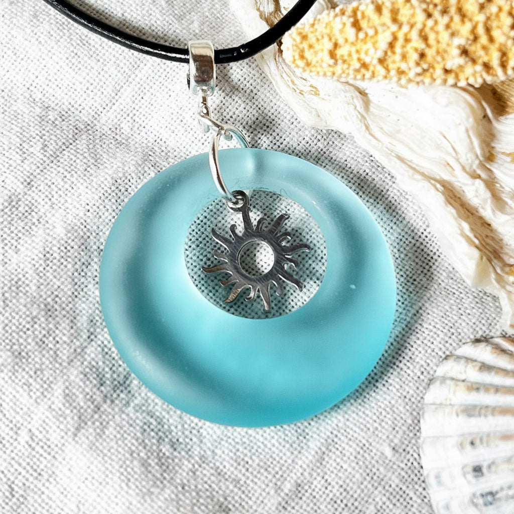 Handgefertigte Halskette mit Donut-Anhänger aus türkisfarbenem Seeglas, kombiniert mit einem filigranen Sonnen-Charm aus Edelstahl. Die Kette besteht aus einer schwarzen Lederschnur (1,5 mm) mit Karabiner und Verlängerungskette aus Edelstahl. Maritimes Schmuckstück für Meerliebhaberinnen und Sommerfreunde. Unikat mit stilvoller Ausstrahlung.