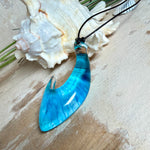 Handgefertigter Haken-Anhänger im Hei-Matau-Stil aus Maserholz und klarem Harz mit Blau-, Türkis- und Weißverlauf, hochglanzpoliert. Oben sichtbares Holz im Harzbereich, getragen an verstellbarem schwarzem Lederband mit Schiebeknoten und Zierperlen. Anhänger ca. 7 × 3 cm, fotografiert mit Muscheln im Hintergrund.