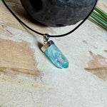 Nahaufnahme einer handgefertigten Halskette mit Aqua Aura Rohkristall: naturbelassener, sechskantiger Bergkristall mit goldbedampfter Aqua-Beschichtung in Türkisblau, gefasst in 925er Silber und getragen an einer schwarzen Lederkette mit Edelstahl-Karabiner und Verlängerungskettchen.