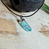 Nahaufnahme einer handgefertigten Halskette mit Aqua Aura Rohkristall: naturbelassener, sechskantiger Bergkristall mit goldbedampfter Aqua-Beschichtung in Türkisblau, gefasst in 925er Silber und getragen an einer schwarzen Lederkette mit Edelstahl-Karabiner und Verlängerungskettchen.