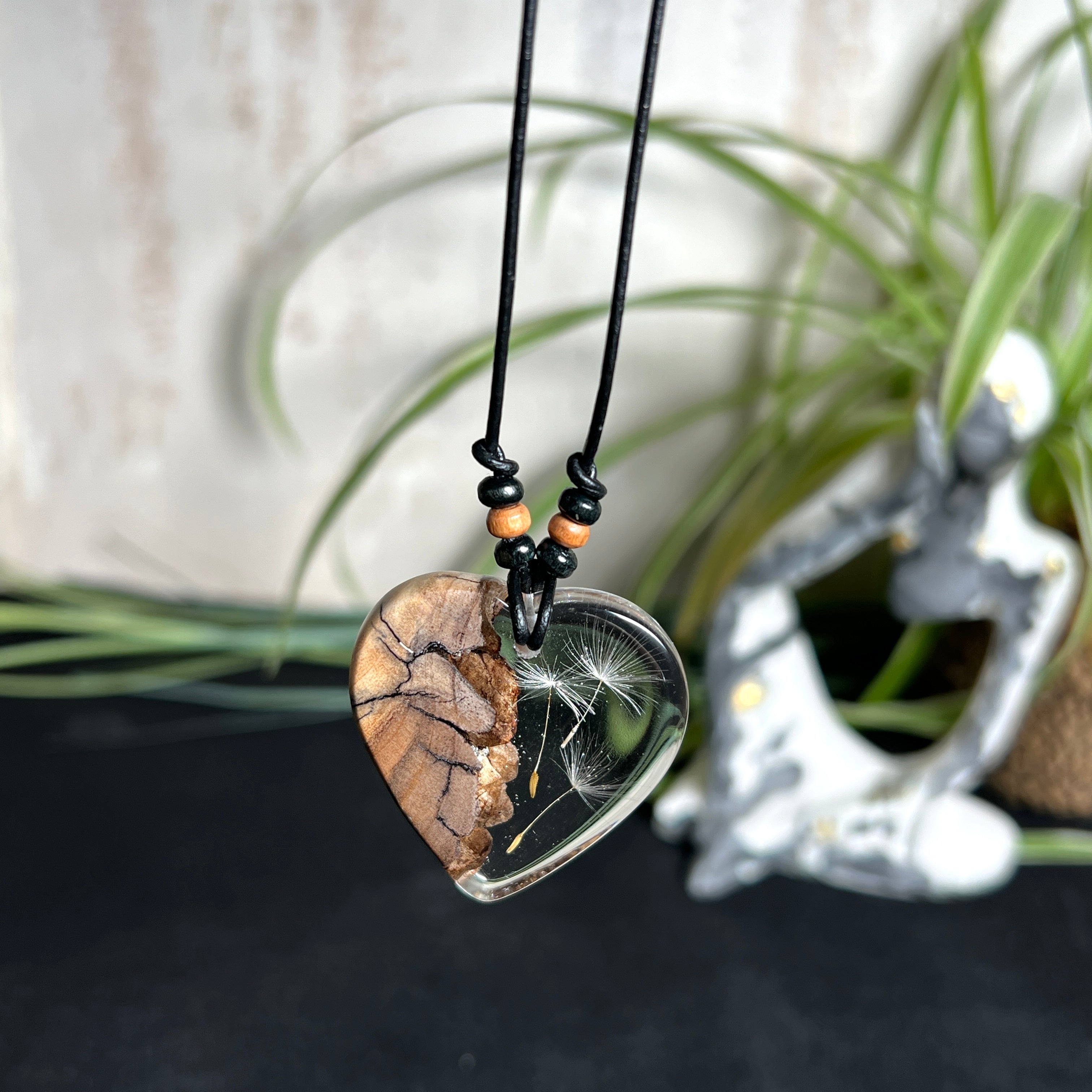 „Handgefertigter Herzanhänger aus Olivenholz und klarem Harz mit echter Pusteblume. Der Anhänger (ca. 3,5 × 3,5 cm) hängt an einem verstellbaren Lederband (23–43 cm) mit Holzperlen. Symbol für Wünsche und Glück, sorgfältig in Handarbeit gegossen, gesägt, geschliffen und poliert. Ein einzigartiger, naturinspirierter Anhänger – ideal als Geschenk oder persönlicher Talisman.“