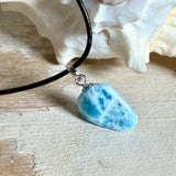 Larimar Freeform-Anhänger auf schwarzer Lederkette. Beidseitig tragbarer Trommelstein, ca. 2 x 1,8 cm groß, mit Silberöse. Der seltene, kostbare Larimar zeigt lebendige Blau- und Weißnuancen wie karibisches Lagunenwasser. Lederkette 45 cm mit Edelstahl-Karabiner und 5 cm Edelstahl-Verlängerung, fotografiert vor einer großen Muschel im maritimen Stil.