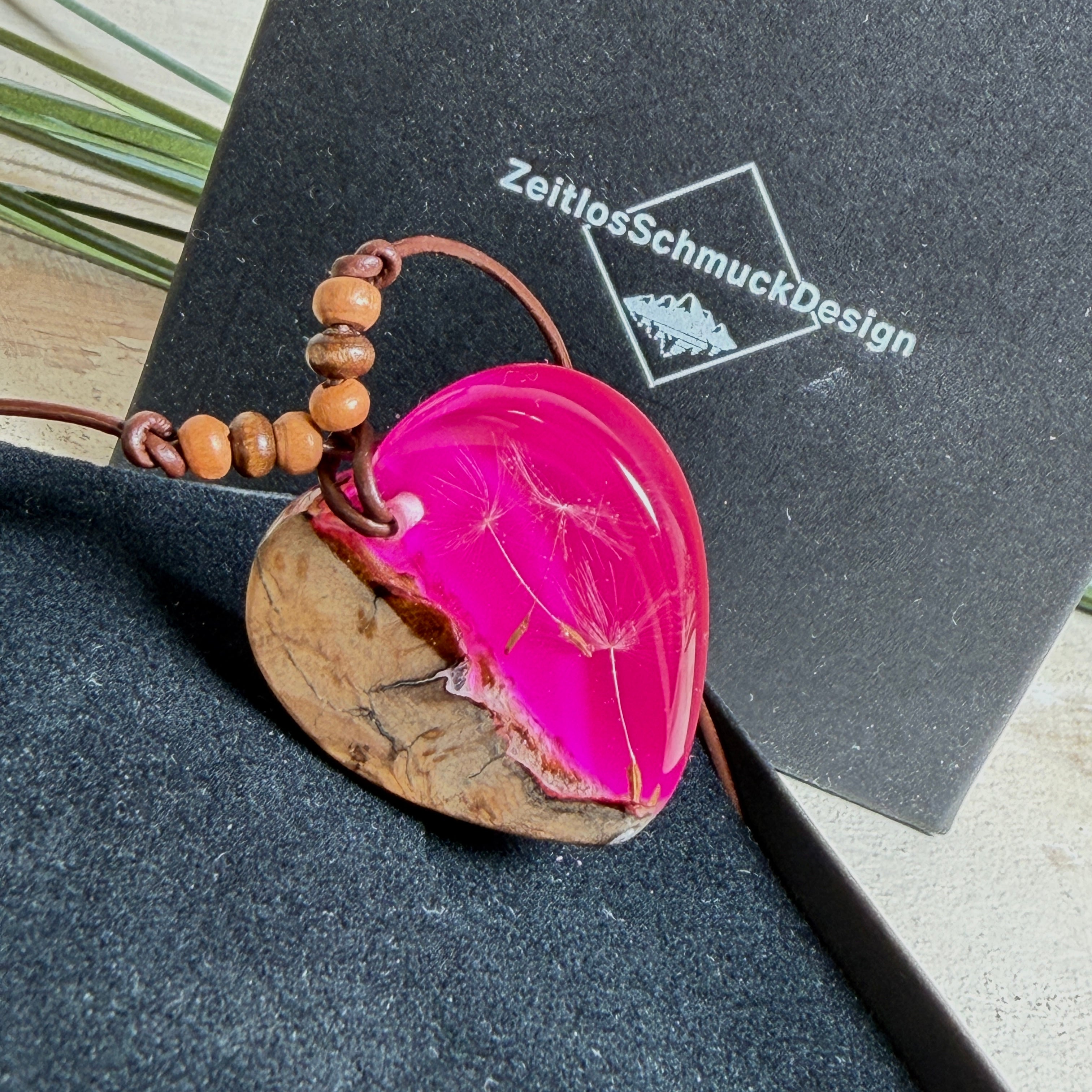 Handgefertigter Herzanhänger aus Olivenholz und leuchtend pinkem Epoxidharz mit eingeschlossenen Pusteblumensamen. Verstellbares braunes Lederband mit Holzperlen, Anhänger ca. 3,5 × 3,3 cm groß. Das Harz leuchtet dezent unter Schwarzlicht. Ein einzigartiges Unikat von ZeitlosSchmuckDesign, inspiriert von Kindheitsglück und Pusteblumen.