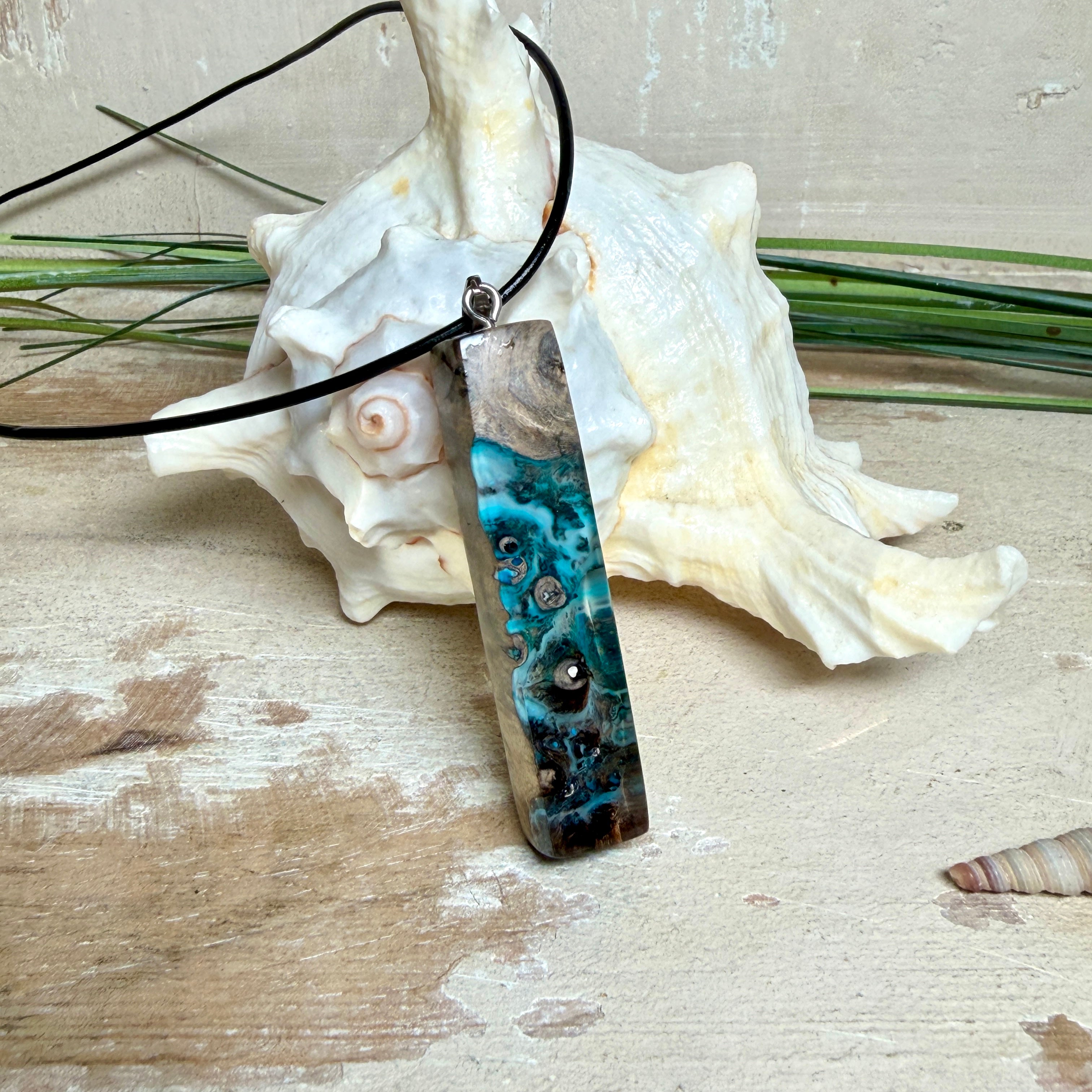 Rechteckiger Stabanhänger aus Maserholz und glasklarem Epoxidharz in tiefen Blau- und Türkistönen mit organischer Lagunen/Riff-Optik. Hochglanzpoliert, ca. 5,3 × 1,2 cm. Der Anhänger hängt an einer schwarzen Lederkette mit Edelstahl-Karabiner und 5-cm-Verlängerung (45 + 5 cm); die Anhängerschlaufe ist ebenfalls aus Edelstahl. Handgefertigtes Unikat, Lieferung in edler Schmuckbox – maritimer Look.