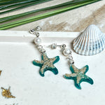 Handgefertigte Seestern-Ohrringe aus Epoxidharz und Sand mit Süßwasserperle, an 925 Sterling Silber Ohrsteckern; versilberte Anhängerschlaufe. Je Seestern ca. 2,6 × 2,5 cm. Leichte, maritime Dangle-Ohrringe im Strand-Look – ideal als Geschenk für Meeresliebhaberinnen.