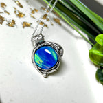 Handgemachter Anhänger mit rundem Cabochon aus Epoxidharz und schimmerndem synthetischem Opal. Die Farben changieren je nach Lichteinfall in Blau, Türkis und Grün. Der Opal sitzt in einer antik versilberten Fassung aus Messing (blei- & nickelfrei). Geliefert mit einer rhodinierten 925 Silberkette (45 cm). Jedes Stück ein Einzelstück – perfekt als stilvolles Geschenk oder persönlicher Glücksbringer.