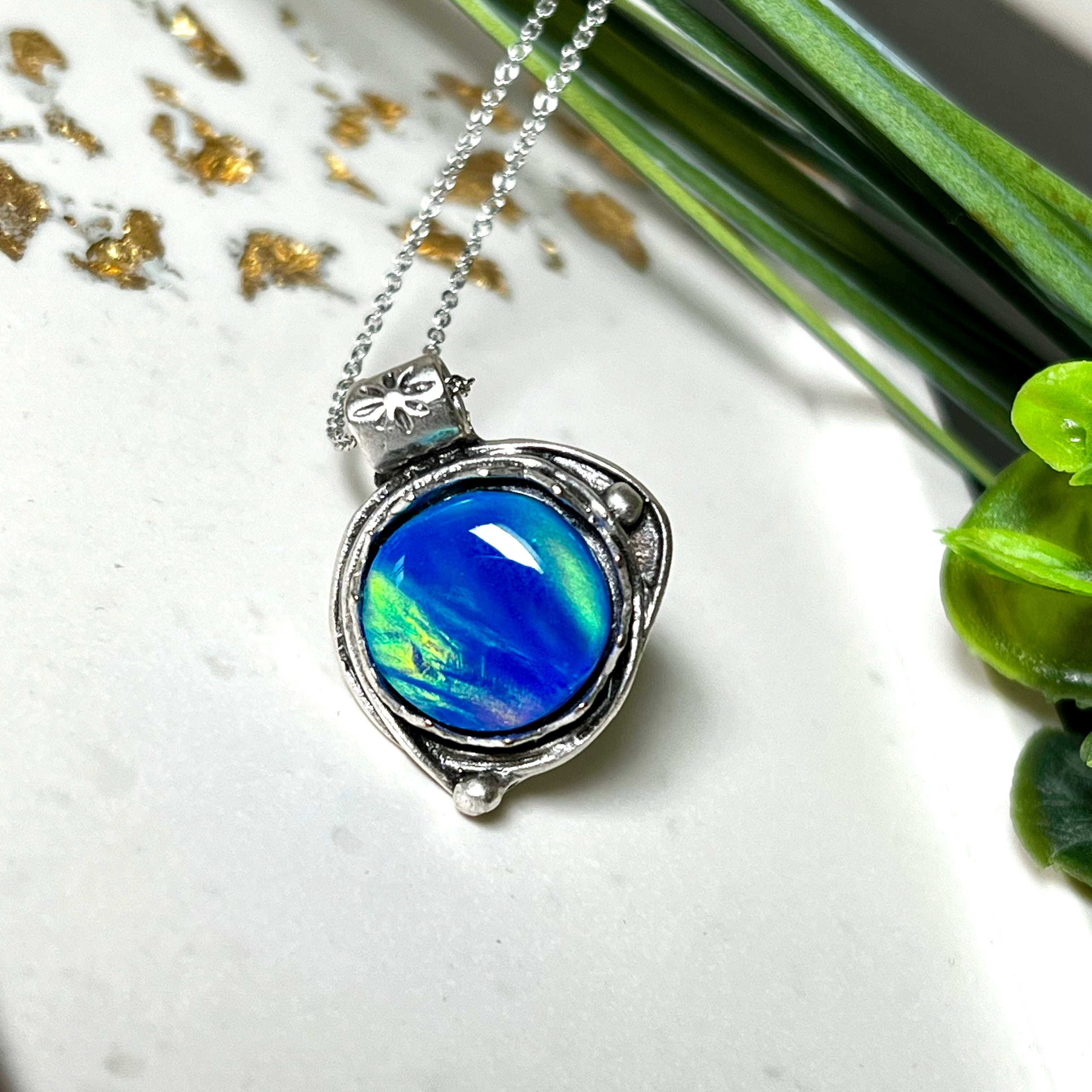 Handgemachter Anhänger mit rundem Cabochon aus Epoxidharz und schimmerndem synthetischem Opal. Die Farben changieren je nach Lichteinfall in Blau, Türkis und Grün. Der Opal sitzt in einer antik versilberten Fassung aus Messing (blei- & nickelfrei). Geliefert mit einer rhodinierten 925 Silberkette (45 cm). Jedes Stück ein Einzelstück – perfekt als stilvolles Geschenk oder persönlicher Glücksbringer.