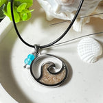 Wave Spirit – runder Edelstahlanhänger mit echter Sandfüllung in Harz eingebettet, inspiriert von einer stilisierten Welle. Verziert mit zwei blauen Achatperlen. Der Anhänger (ca. 2,5 cm Durchmesser) hängt an einer 45 cm langen Lederkette mit Edelstahlverschluss und 5 cm Verlängerungskette. Maritimer Look trifft moderne Eleganz – ein handgefertigtes Schmuckstück für Meerliebhaber