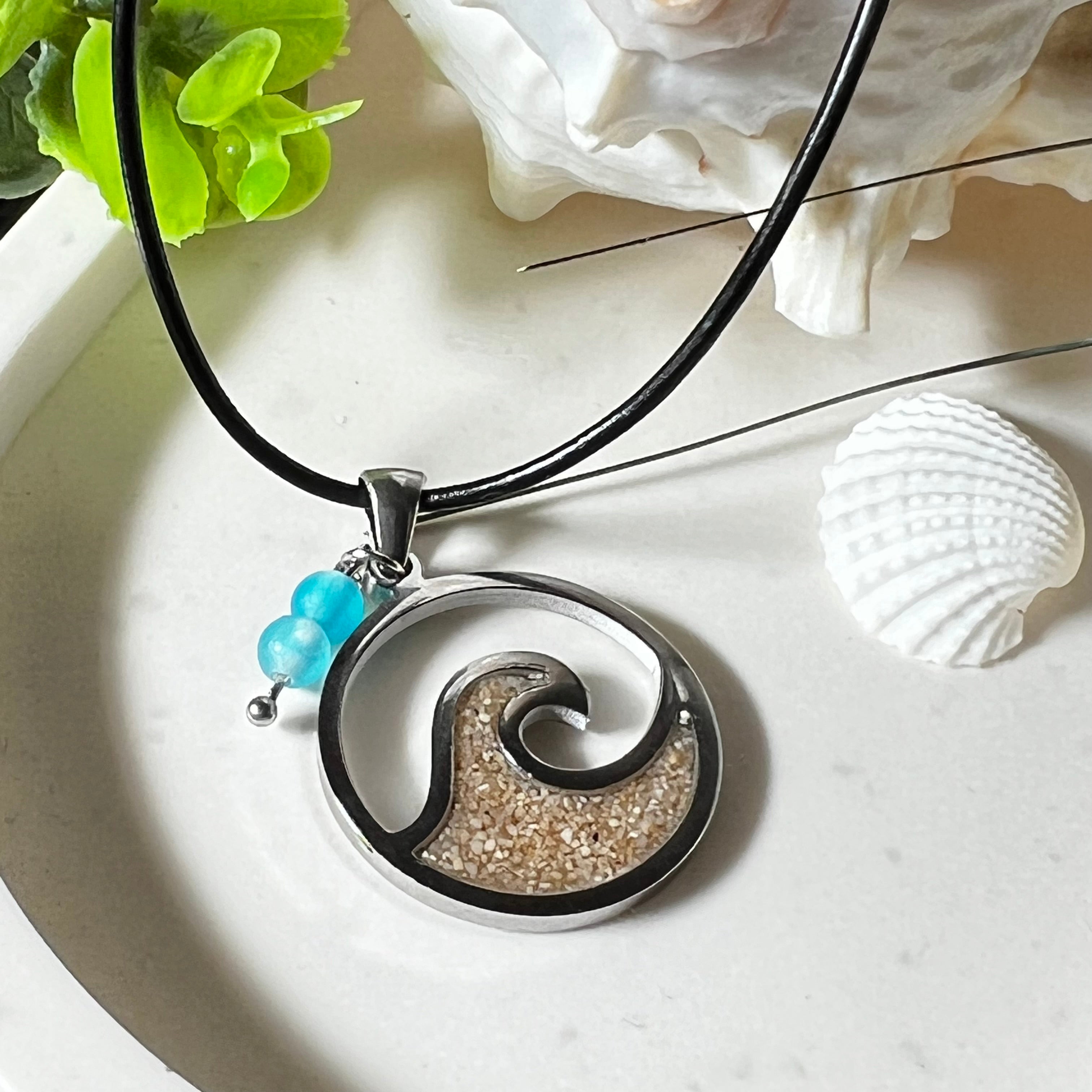 Wave Spirit – runder Edelstahlanhänger mit echter Sandfüllung in Harz eingebettet, inspiriert von einer stilisierten Welle. Verziert mit zwei blauen Achatperlen. Der Anhänger (ca. 2,5 cm Durchmesser) hängt an einer 45 cm langen Lederkette mit Edelstahlverschluss und 5 cm Verlängerungskette. Maritimer Look trifft moderne Eleganz – ein handgefertigtes Schmuckstück für Meerliebhaber
