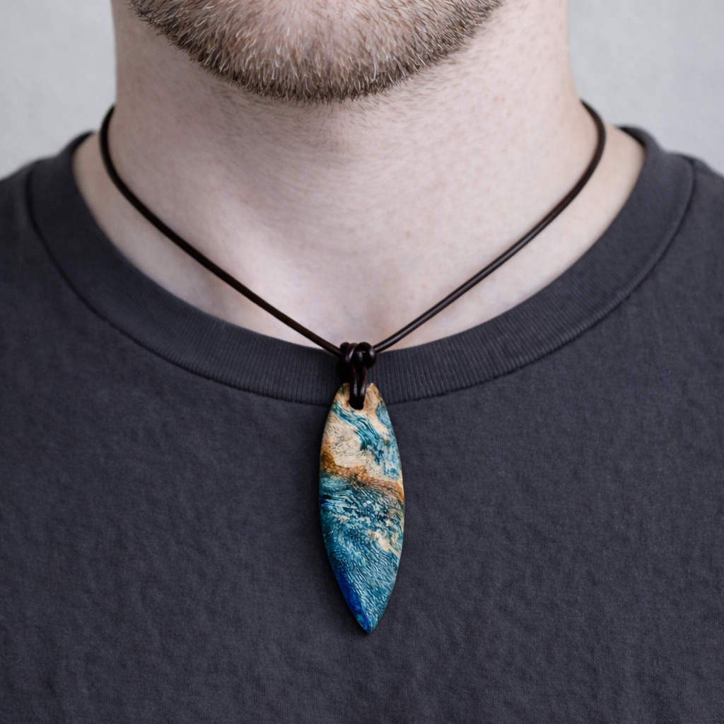 Unikat Anhänger aus stabilisiertem Holz mit türkisblauem Harzverlauf, schlanke Form, poliert. Verstellbares braunes Lederband 22–46 cm mit Schiebeknoten. Anhänger ca. 6,6 × 2 cm. Tragefoto am Hals und Produktfotos mit maritimer Deko