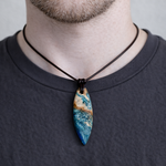 Unikat Anhänger aus stabilisiertem Holz mit türkisblauem Harzverlauf, schlanke Form, poliert. Verstellbares braunes Lederband 22–46 cm mit Schiebeknoten. Anhänger ca. 6,6 × 2 cm. Tragefoto am Hals und Produktfotos mit maritimer Deko