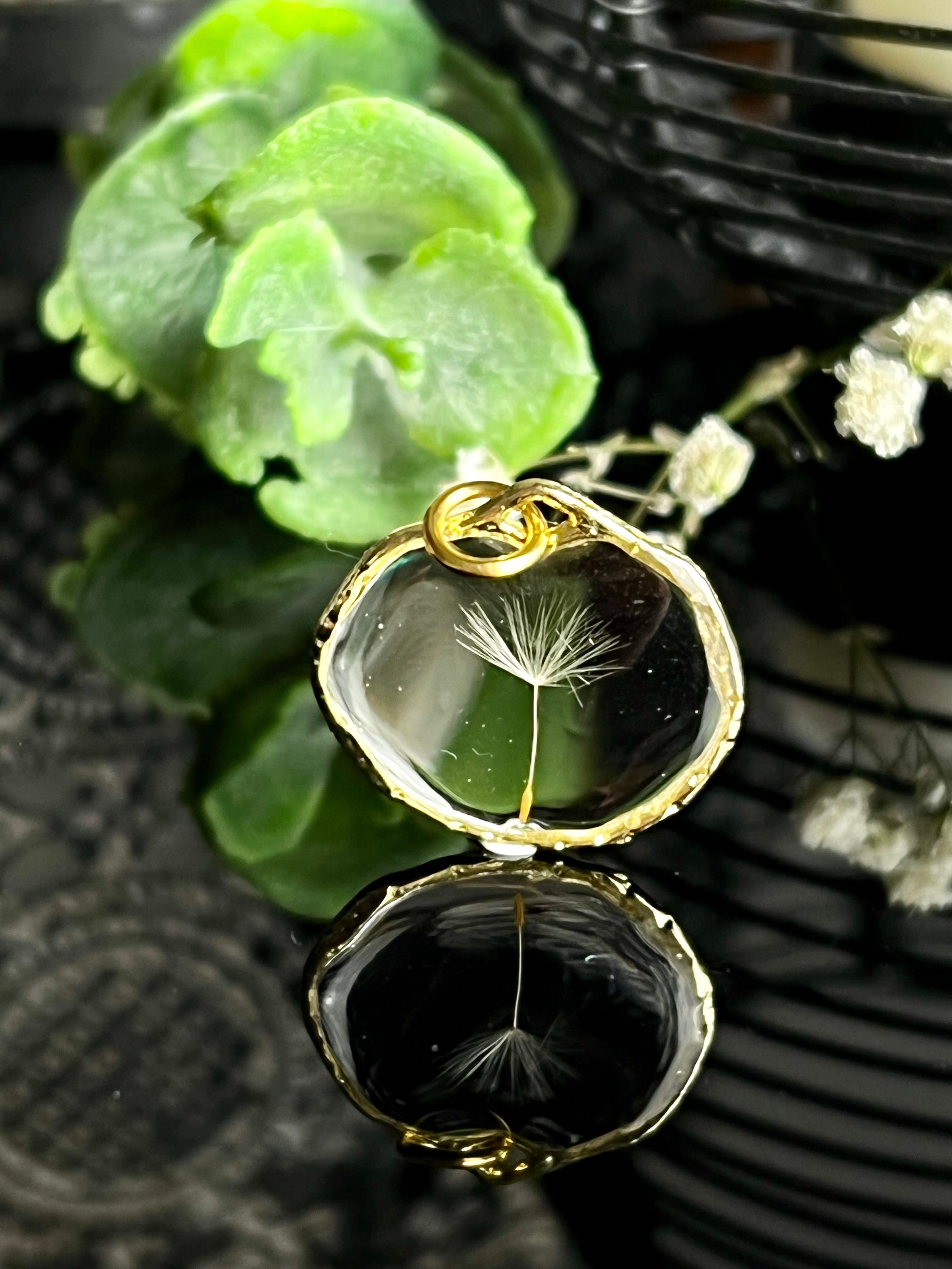 “Eleganter goldener Anhänger mit einer einzelnen Pusteblumen-Samenkapsel, eingefasst in transparentem Harz. Präsentiert auf einem reflektierenden Untergrund mit natürlichen grünen Blättern im Hintergrund. Perfektes Accessoire für Liebhaber zeitloser und natürlicher Schmuckdesigns.”