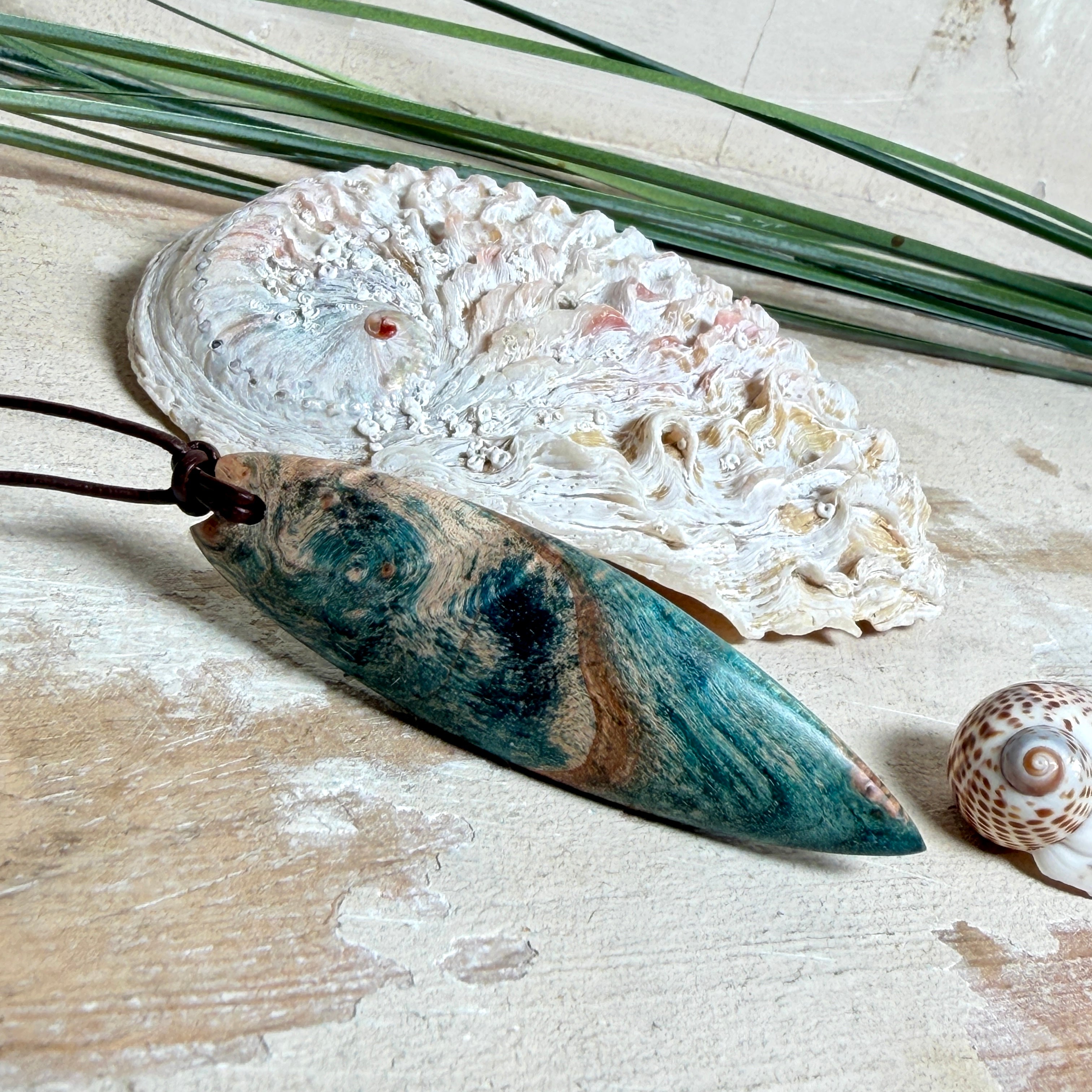 Unikat Anhänger aus stabilisiertem Holz mit türkisblauem Harzverlauf, schlanke Form, poliert. Verstellbares braunes Lederband 22–46 cm mit Schiebeknoten. Anhänger ca. 6,6 × 2 cm. Tragefoto am Hals und Produktfotos mit maritimer Deko