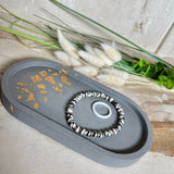 Handgefertigtes ovales Schmucktablett aus grauen  Jesmonite mit schimmernden Blattgold-Details. Die Unterseite ist mit vier Korkpunkten versehen und schützt empfindliche Oberflächen. Ideal zur stilvollen Aufbewahrung von Schmuck, Schlüsseln, Brille oder kleinen Dekoartikeln. Jedes Tablett ist ein Unikat mit individueller Maserung und goldenen Akzenten – Lieferung ohne Deko. Die 