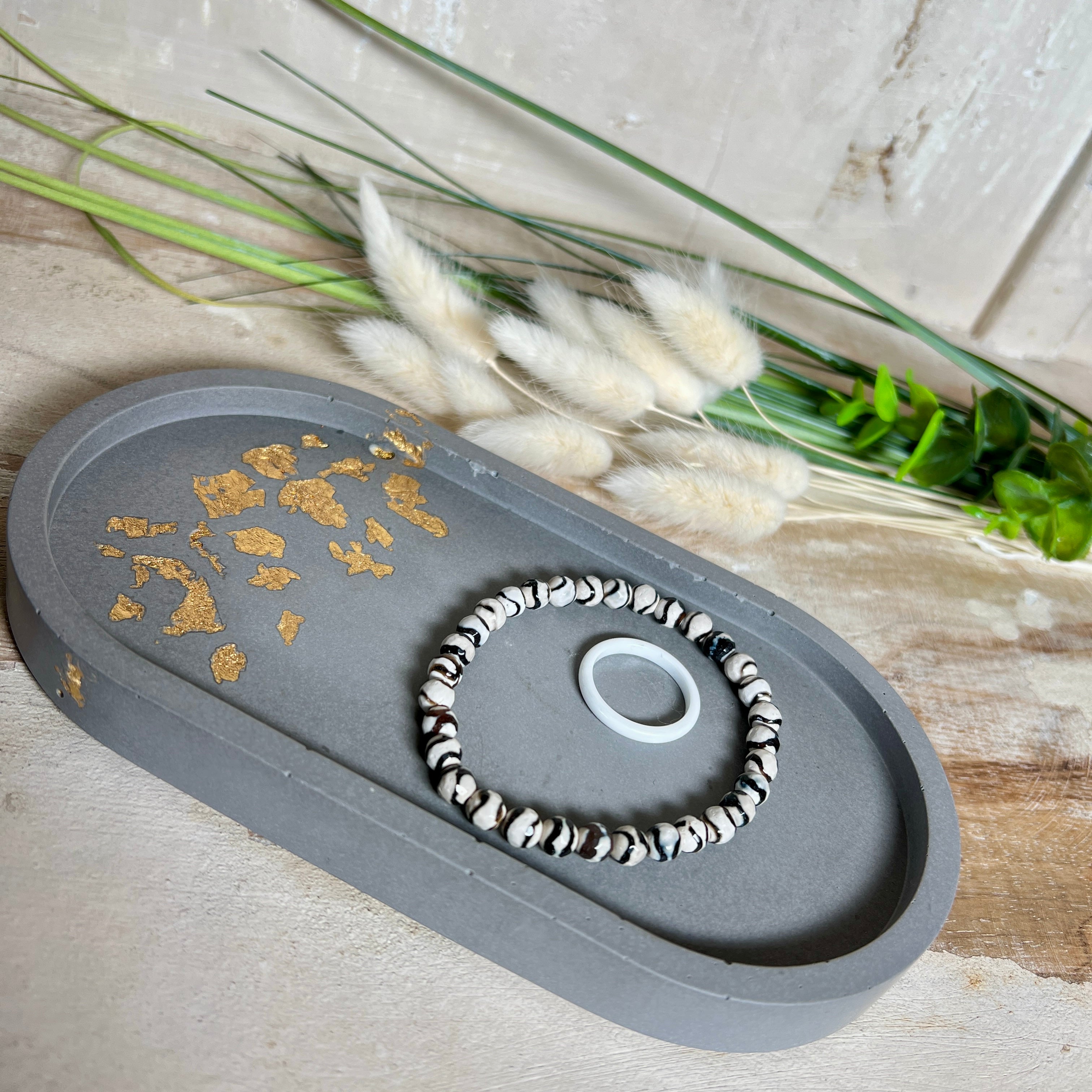 Handgefertigtes ovales Schmucktablett aus grauen  Jesmonite mit schimmernden Blattgold-Details. Die Unterseite ist mit vier Korkpunkten versehen und schützt empfindliche Oberflächen. Ideal zur stilvollen Aufbewahrung von Schmuck, Schlüsseln, Brille oder kleinen Dekoartikeln. Jedes Tablett ist ein Unikat mit individueller Maserung und goldenen Akzenten – Lieferung ohne Deko. Die 