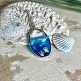 „Riffschatz“ – ovaler Harz-Anhänger mit plastischer 3D-Welle in Blau/Türkis, weißer Schaumkrone und Muschel-/Korallen-Stickern am Grund. Die Sticker sind unter klarem Harz versiegelt. Silber-Legierung, ca. 3,9 × 2,3 cm, an einer ca. 56 cm langen versilberten Kette. Handgefertigt, hochglänzend, jedes Stück ein Unikat.