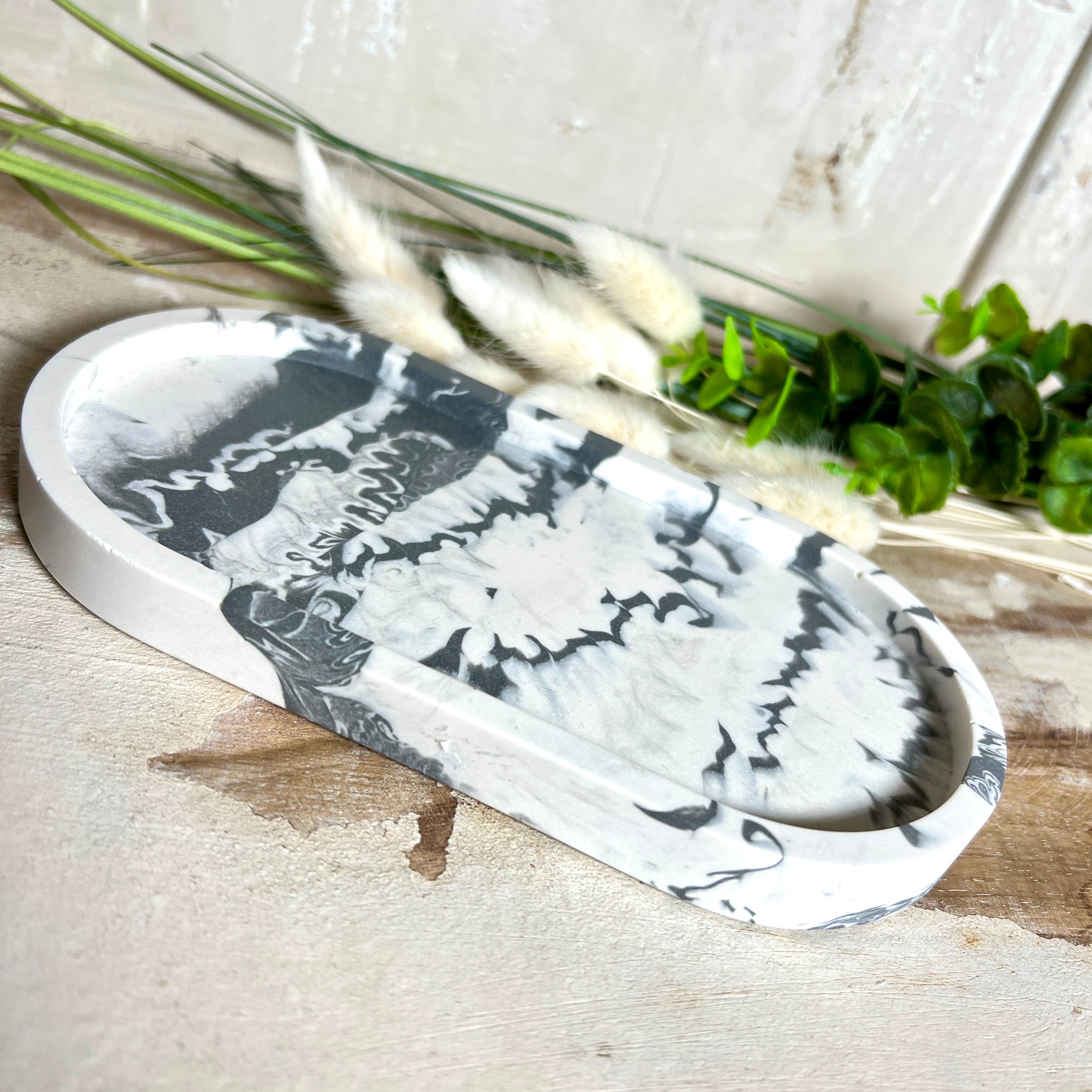 Handgegossenes Schmucktablett aus Jesmonite mit grau -weißer Marmorierung. Die ovale Form und die individuelle Musterung machen jedes Stück einzigartig. Die Rückseite ist mit rutschfesten Korkpunkten ausgestattet – ideal als stilvolle Ablage für Schmuck, Kristalle, Schlüssel oder Brille. Unikat in modernem Monochrom-Design. Lieferung ohne Deko.