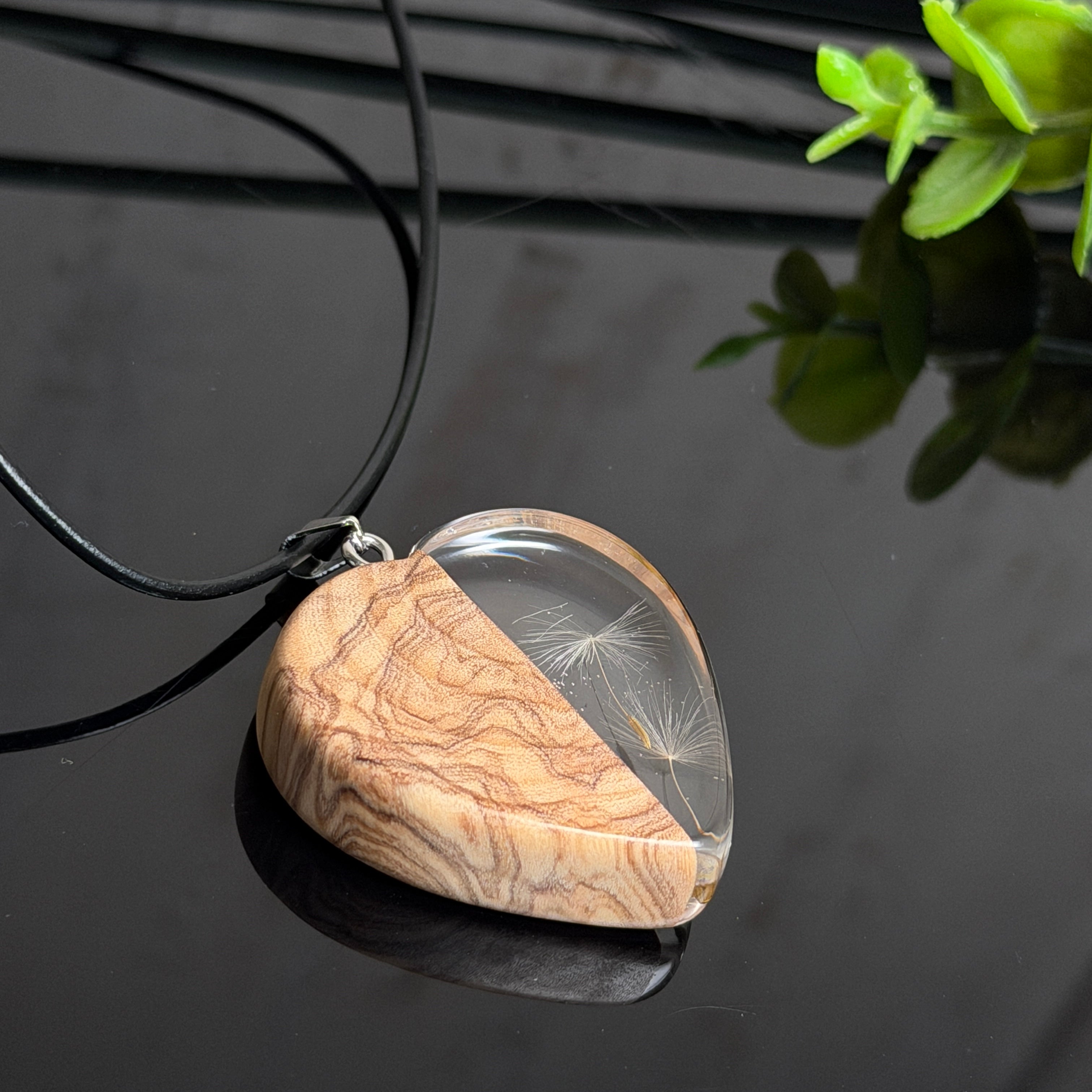 Pusteblumen Herzanhänger aus Olivenholz und glasklarem Epoxidharz, echte Löwenzahnsamen im Harz schwebend, schwarze Lederkette, handgefertigter Naturschmuck mit Spiegelung, dunkler Hintergrund