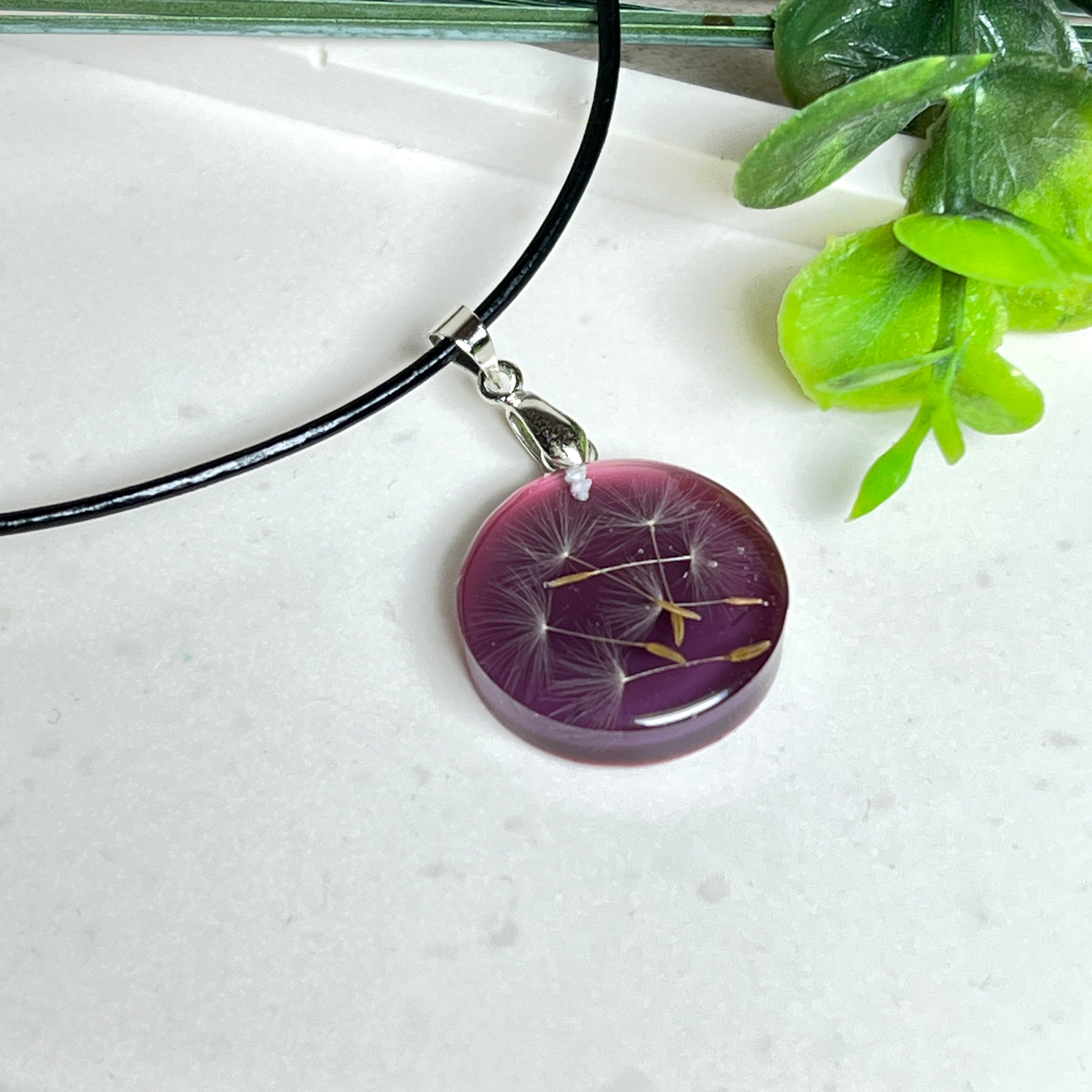 Runder Pusteblumen-Anhänger aus Harz, Hintergrund Brombeerfarben. Durchmesser 22 mm, Edelstahlöse, Lederkette verstellbar mit Edelstahlverschluss.