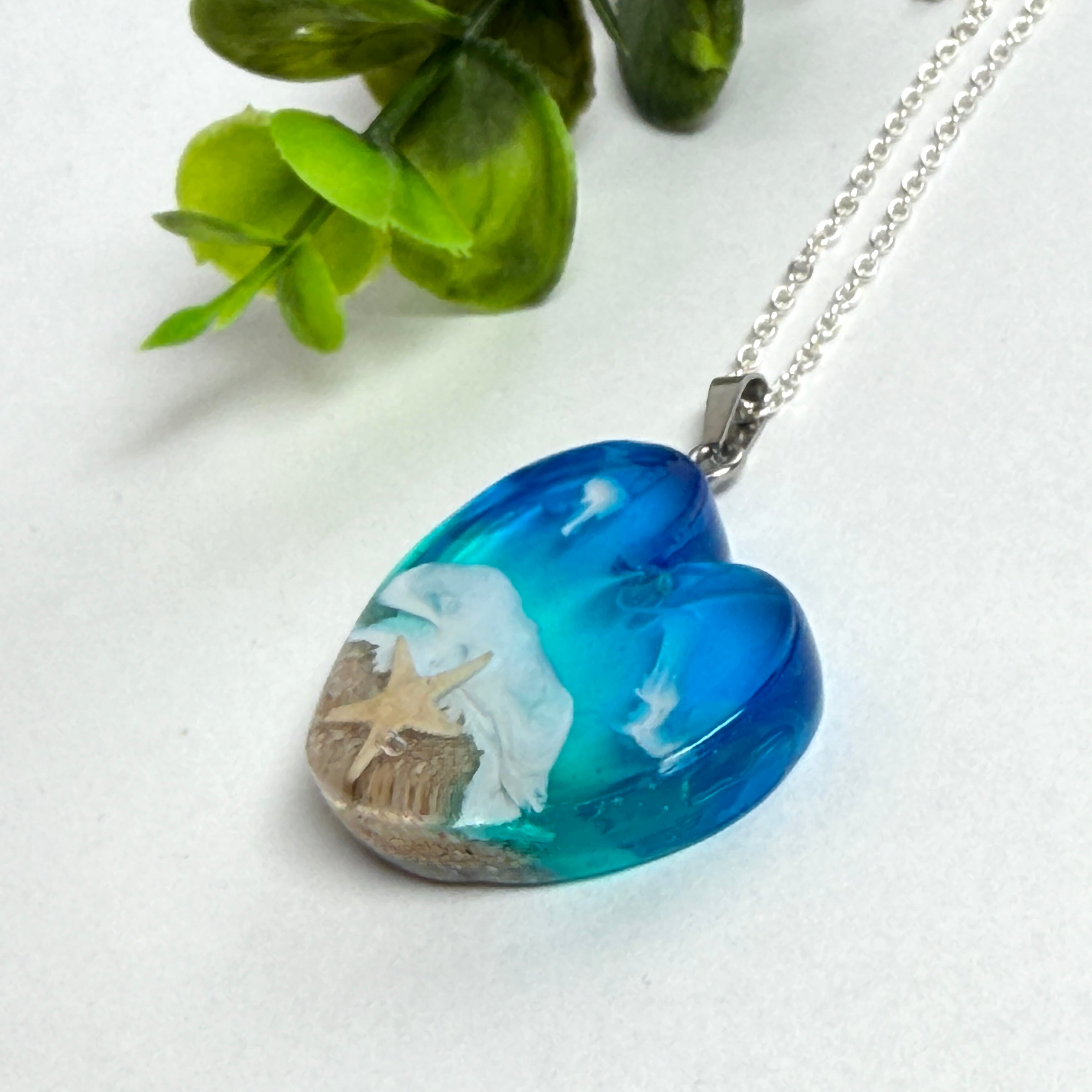 Ein herzförmiger Anhänger mit einer ganzen Ozeanwelt darin: Welle, Sand und Seestern in leuchtendem Blau und Türkis – handgegossen, einzigartig, unvergesslich. Mit versilberter Kette (46 cm) und Edelstahl-Karabiner.