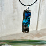 Rechteckiger Stabanhänger aus Maserholz und glasklarem Epoxidharz in tiefen Blau- und Türkistönen mit organischer Lagunen/Riff-Optik. Hochglanzpoliert, ca. 4,6 × 1,2 cm. Der Anhänger hängt an einer schwarzen Lederkette mit Edelstahl-Karabiner und 5-cm-Verlängerung (45 + 5 cm); die Anhängerschlaufe ist ebenfalls aus Edelstahl. Handgefertigtes Unikat, Lieferung in edler Schmuckbox – maritimer Look.