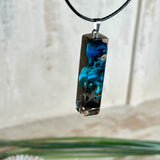 Rechteckiger Stabanhänger aus Maserholz und glasklarem Epoxidharz in tiefen Blau- und Türkistönen mit organischer Lagunen/Riff-Optik. Hochglanzpoliert, ca. 4,6 × 1,2 cm. Der Anhänger hängt an einer schwarzen Lederkette mit Edelstahl-Karabiner und 5-cm-Verlängerung (45 + 5 cm); die Anhängerschlaufe ist ebenfalls aus Edelstahl. Handgefertigtes Unikat, Lieferung in edler Schmuckbox – maritimer Look.