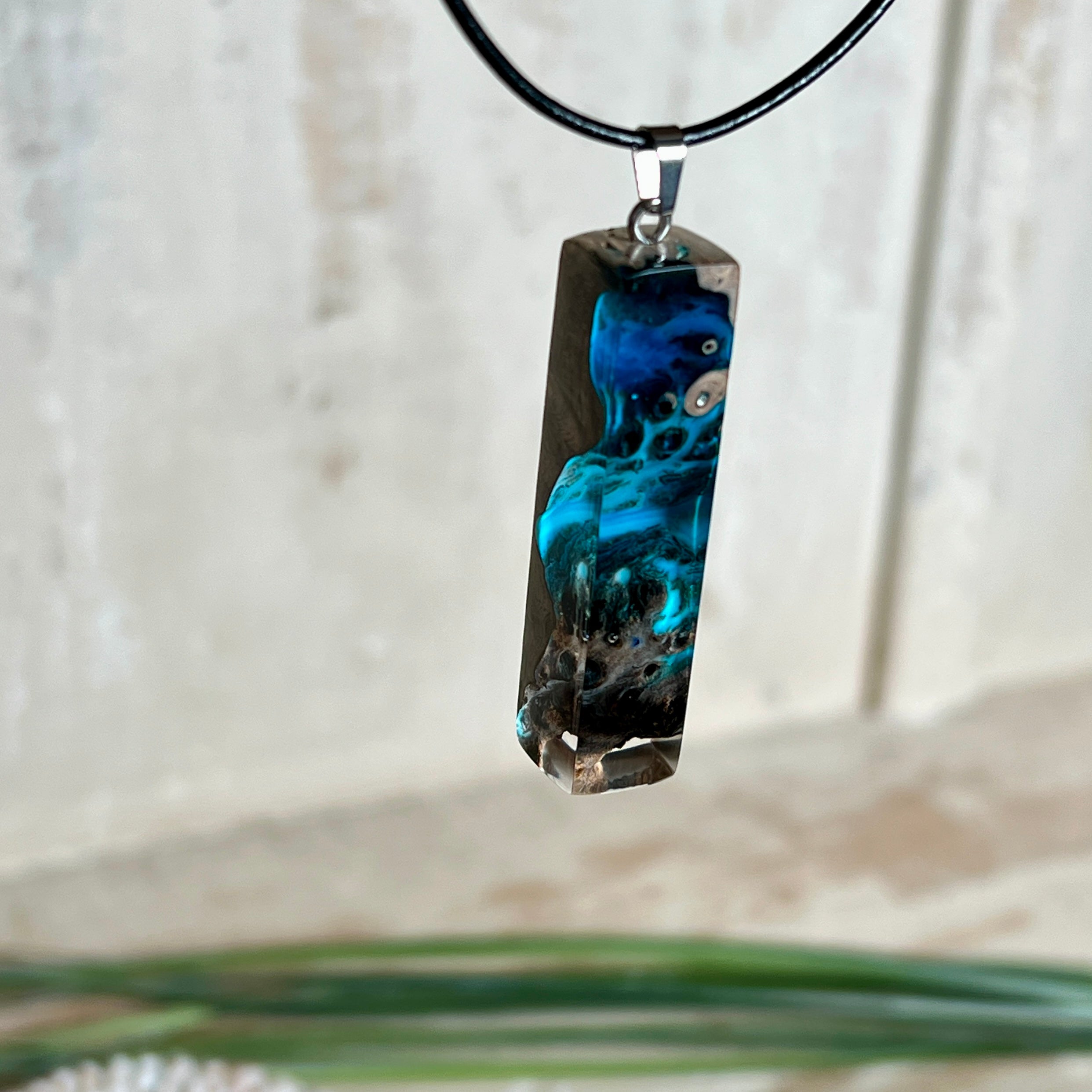 Rechteckiger Stabanhänger aus Maserholz und glasklarem Epoxidharz in tiefen Blau- und Türkistönen mit organischer Lagunen/Riff-Optik. Hochglanzpoliert, ca. 4,6 × 1,2 cm. Der Anhänger hängt an einer schwarzen Lederkette mit Edelstahl-Karabiner und 5-cm-Verlängerung (45 + 5 cm); die Anhängerschlaufe ist ebenfalls aus Edelstahl. Handgefertigtes Unikat, Lieferung in edler Schmuckbox – maritimer Look.