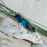 Rechteckiger Stabanhänger aus Maserholz und glasklarem Epoxidharz in tiefen Blau- und Türkistönen mit organischer Lagunen/Riff-Optik. Hochglanzpoliert, ca. 4,6 × 1,2 cm. Der Anhänger hängt an einer schwarzen Lederkette mit Edelstahl-Karabiner und 5-cm-Verlängerung (45 + 5 cm); die Anhängerschlaufe ist ebenfalls aus Edelstahl. Handgefertigtes Unikat, Lieferung in edler Schmuckbox – maritimer Look.