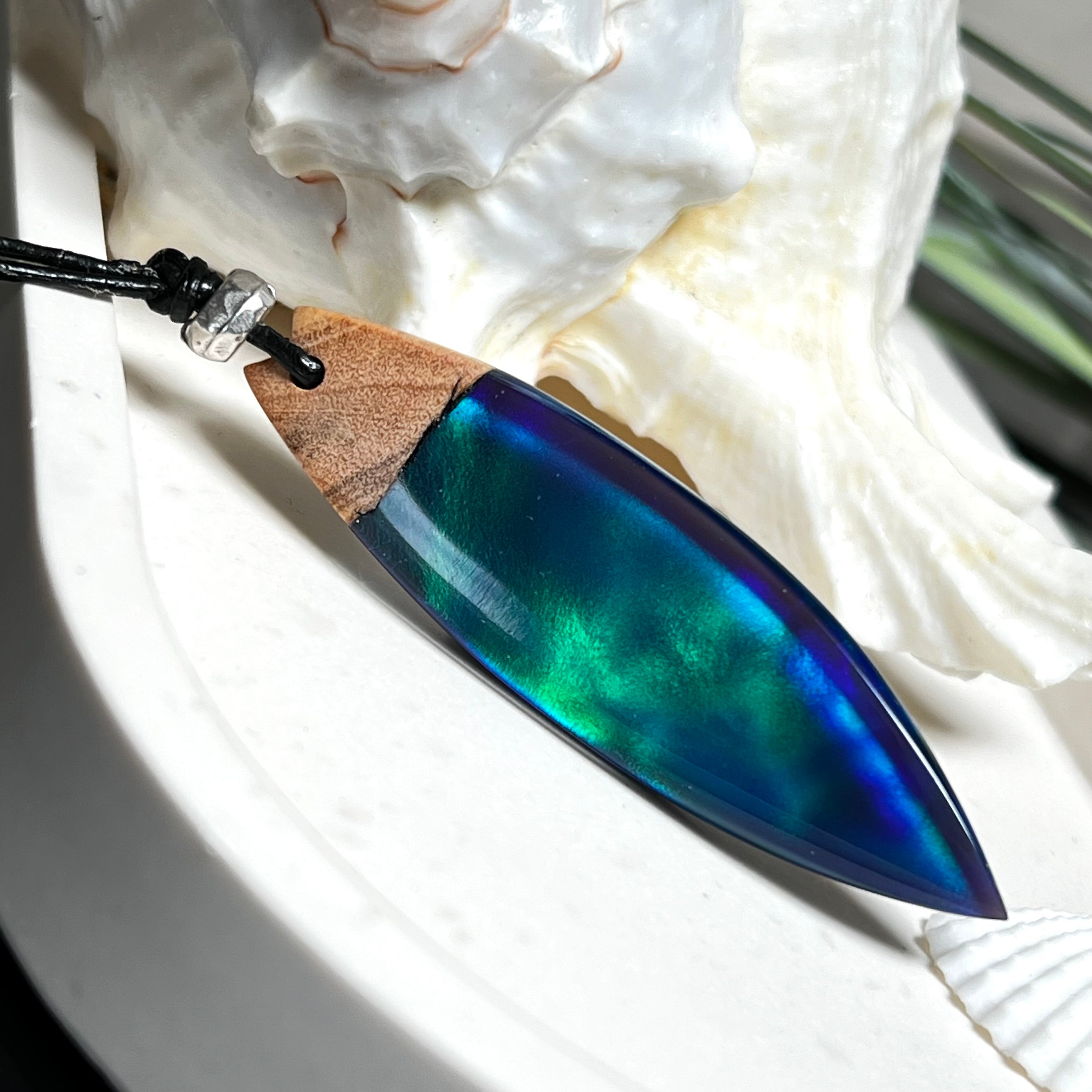 Beidseitig tragbarer Surfbrett-Anhänger aus Olivenholz, kombiniert mit klaren Epoxidharz in Blau-Grün. Eingearbeiteter synthetischer Opal erzeugt ein faszinierendes Farbspiel wie Nordlichter. Größe ca. 6 × 1,8 cm. Mit verstellbarem Lederband (26–45 cm), dekorativen Perlen & Schiebeknoten. Handgefertigt & einzigartig. Ein Schmuckstück mit Naturinspiration.