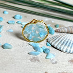 Runder Anhänger „Golden Atlantis Dream“ mit kleinen Larimar-Edelsteinchen in klarem Resin, gefasst in einer 24K vergoldeten Zinklegierung in Shiny Gold. Die Larimarsteine zeigen sanfte Blau- und Weißtöne wie karibische Wellen. Durchmesser ca. 22 mm, handgefertigtes Unikat, fotografiert mit Larimarsteinchen, Muscheln und einer 2-Euro-Münze zur Größenorientierung auf maritimem Untergrund.