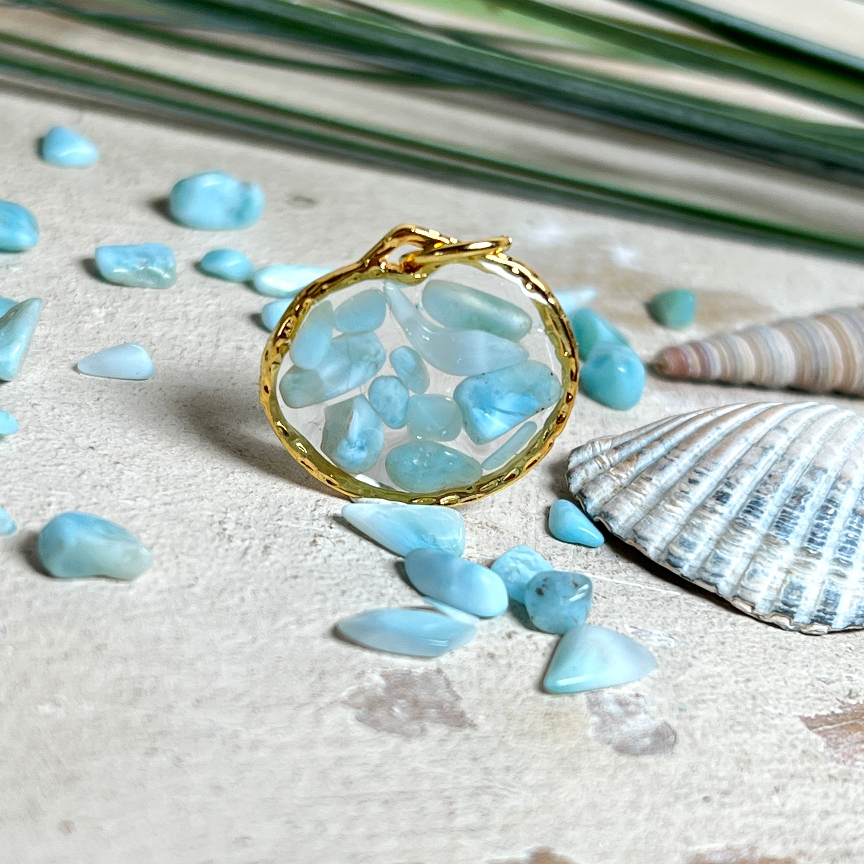 Runder Anhänger „Golden Atlantis Dream“ mit kleinen Larimar-Edelsteinchen in klarem Resin, gefasst in einer 24K vergoldeten Zinklegierung in Shiny Gold. Die Larimarsteine zeigen sanfte Blau- und Weißtöne wie karibische Wellen. Durchmesser ca. 22 mm, handgefertigtes Unikat, fotografiert mit Larimarsteinchen, Muscheln und einer 2-Euro-Münze zur Größenorientierung auf maritimem Untergrund.