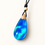 Cleanes Hero-Bild eines handgefertigten Tropfenanhängers aus Maserholz, blau-grün schimmerndem Epoxidharz mit Labor-Opal-Effekt und schwarzem Lederband mit Holzperle vor hellem Hintergrund.