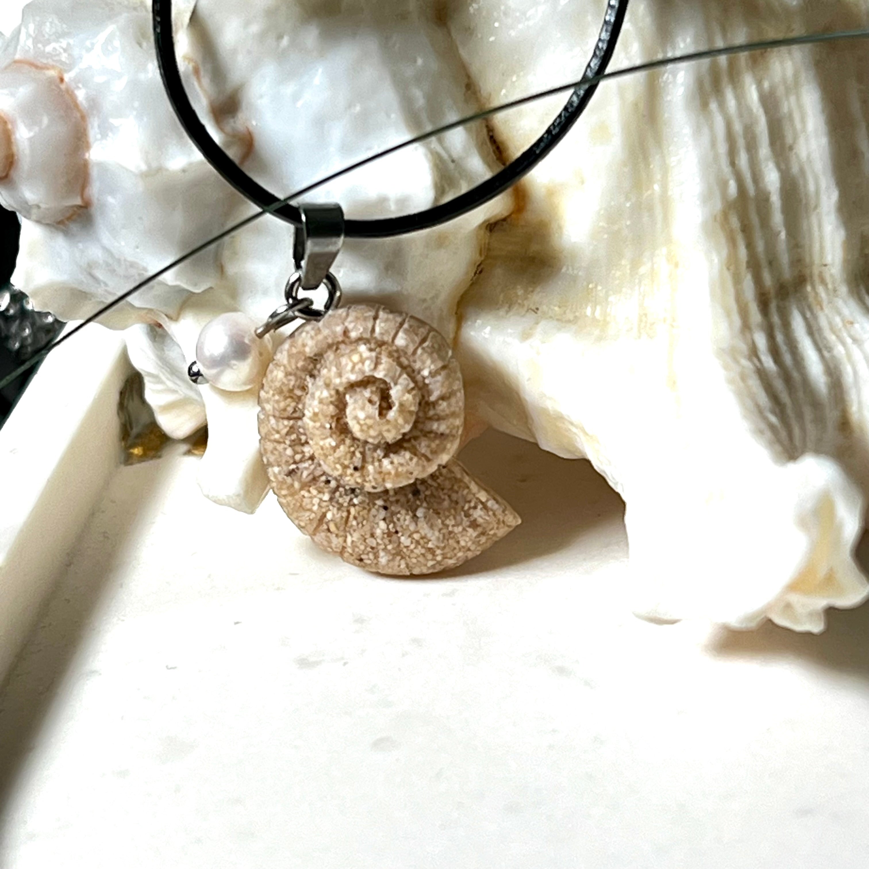 Handgefertigter Anhänger in Ammonit-Form aus echtem Meersand und Harz, kombiniert mit einer weißen Süßwasserperle. Der maritime Anhänger hängt an einer schwarzen Lederkette mit Edelstahlöse und Karabinerverschluss. Im Hintergrund liegt eine weiße Muschel, die das natürliche, strandinspirierte Design des Schmuckstücks unterstreicht. Ideal als besonderes Geschenk oder Erinnerung an den Sommer.
