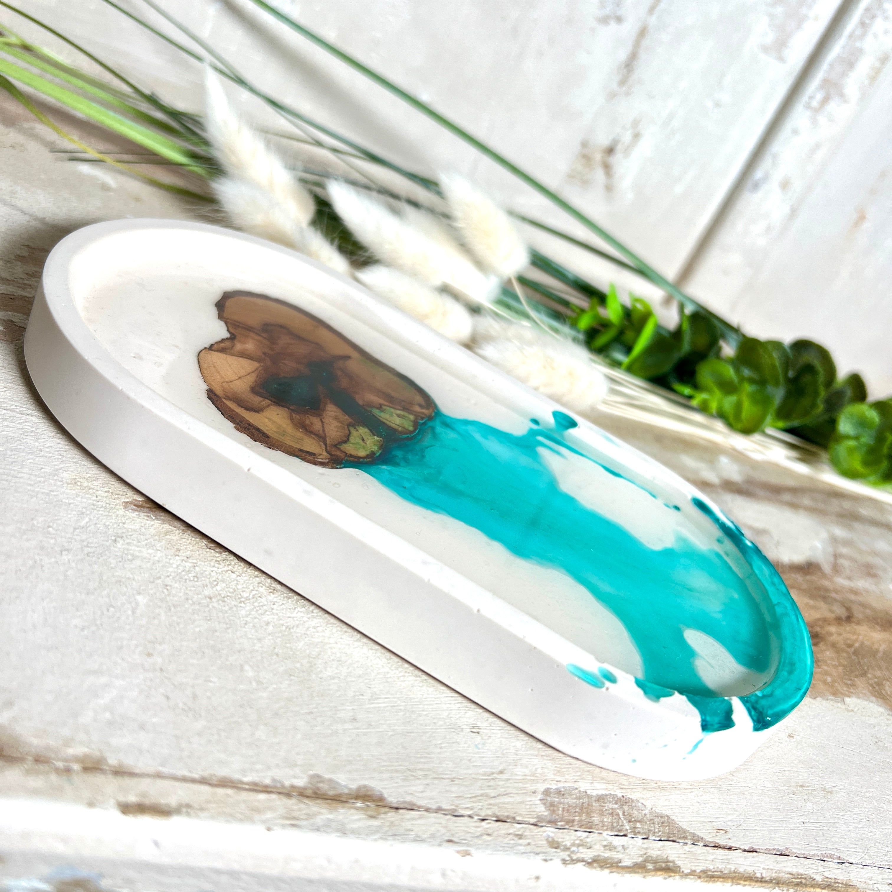 Handgefertigtes Schmucktablett aus Jesmonite mit eingebettetem Olivenholz und türkisfarbenem Resin. Die glänzende Oberfläche erinnert an Wasserstrukturen, während das Naturholz Tiefe und Wärme verleiht. Die Rückseite ist mit Korkpunkten versehen und schützt empfindliche Oberflächen. Perfekt als Ablage für Schmuck, Brille, Schlüssel oder Deko jedes Stück ein Unikat.