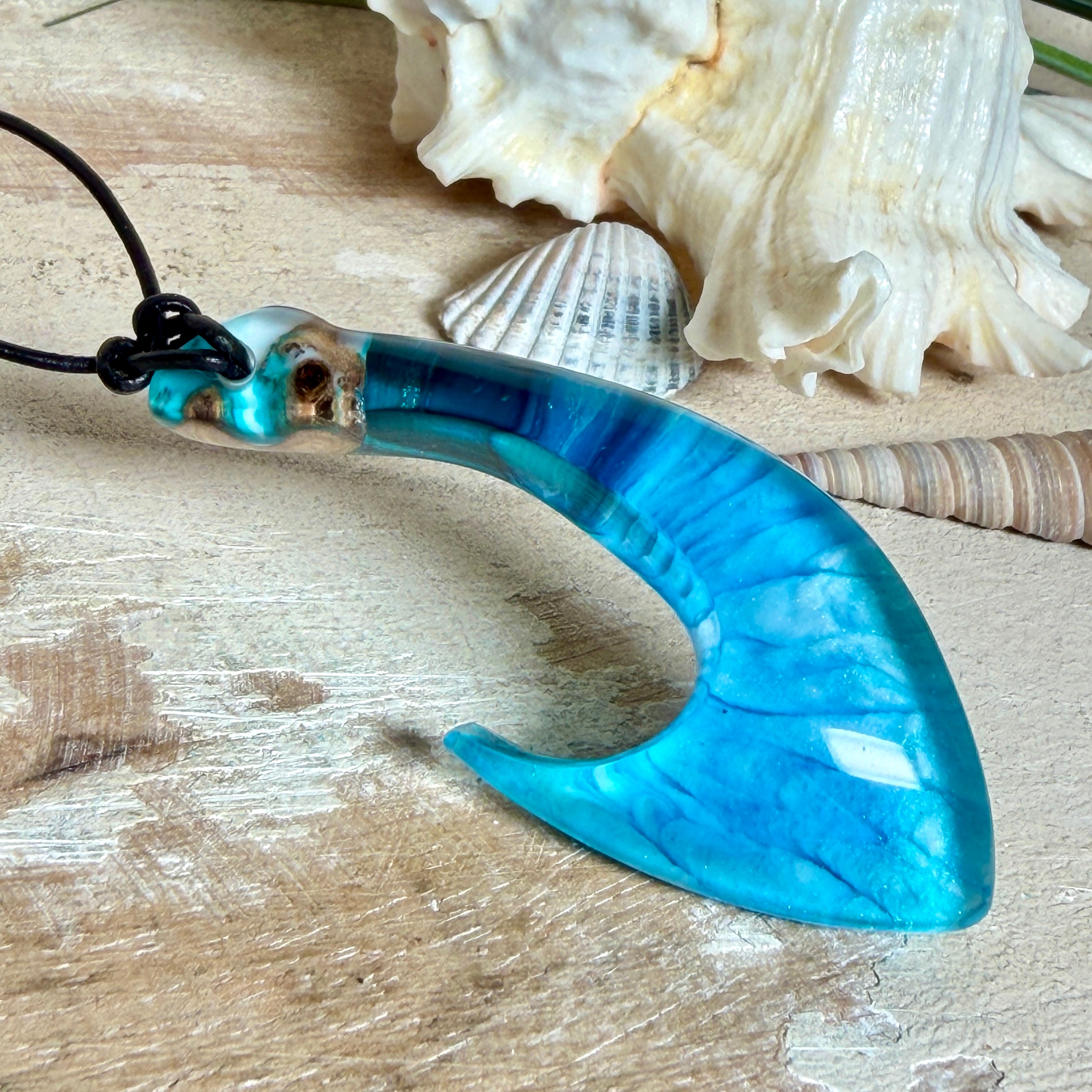 Handgefertigter Haken-Anhänger im Hei-Matau-Stil aus Maserholz und klarem Harz mit Blau-, Türkis- und Weißverlauf, hochglanzpoliert. Oben sichtbares Holz im Harzbereich, getragen an verstellbarem schwarzem Lederband mit Schiebeknoten und Zierperlen. Anhänger ca. 7 × 3 cm, fotografiert mit Muscheln im Hintergrund.