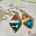 Handgefertigte Ohrringe „Ocean Diamond“ aus Olivenholz und Epoxidharz in Diamantform. Türkis-, Blau- und Weißtöne bilden ein wellenartiges Design, inspiriert vom Meer. Jeder Ohrring ist ein Unikat mit individueller Maserung. Vergoldete, nickelfreie und bleifreie Anhängerschlaufe, 925 Sterling Silber Ohrhaken. Maritimer Schmuck für Meerliebhaber und besondere Anlässe.