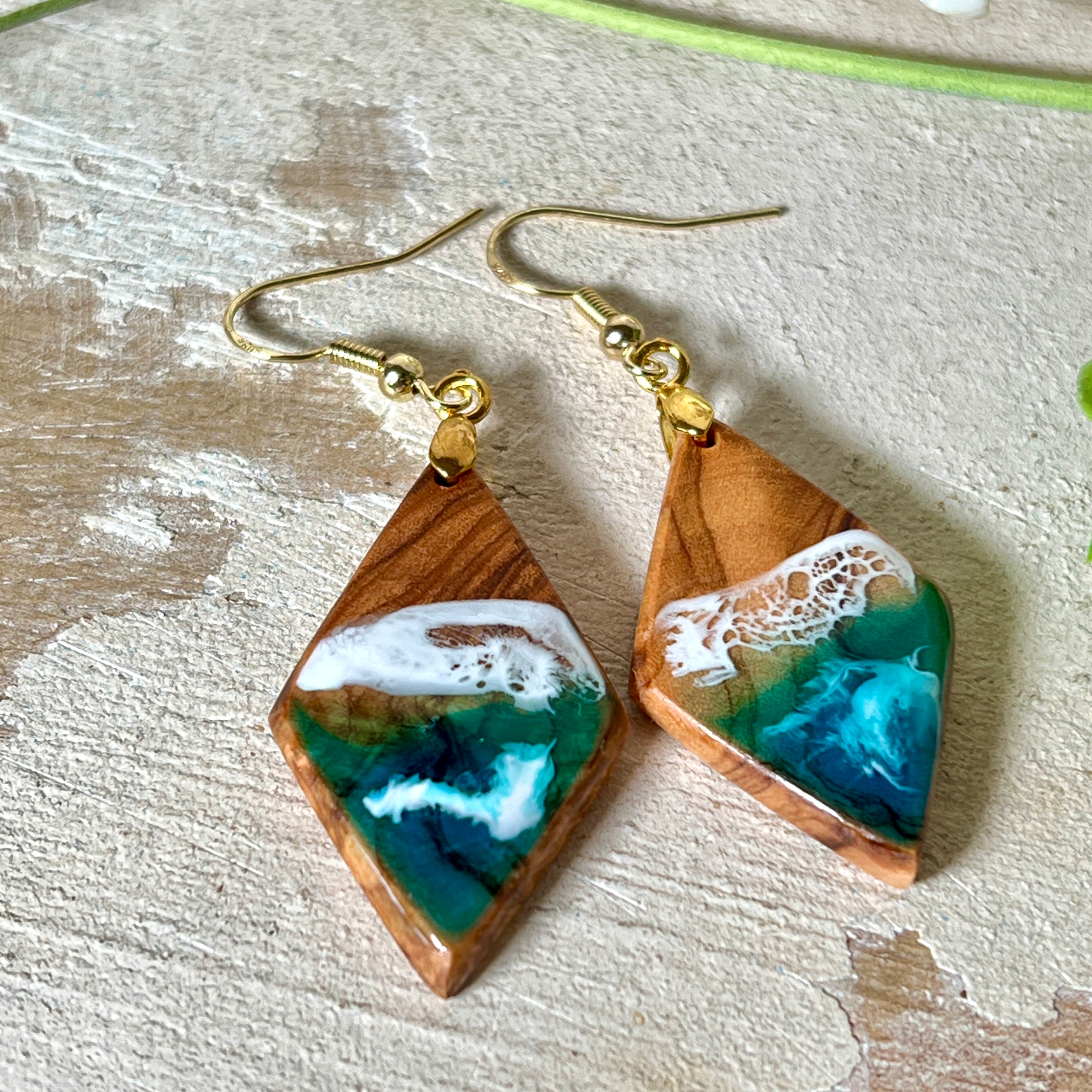 Handgefertigte Ohrringe „Ocean Diamond“ aus Olivenholz und Epoxidharz in Diamantform. Türkis-, Blau- und Weißtöne bilden ein wellenartiges Design, inspiriert vom Meer. Jeder Ohrring ist ein Unikat mit individueller Maserung. Vergoldete, nickelfreie und bleifreie Anhängerschlaufe, 925 Sterling Silber Ohrhaken. Maritimer Schmuck für Meerliebhaber und besondere Anlässe.